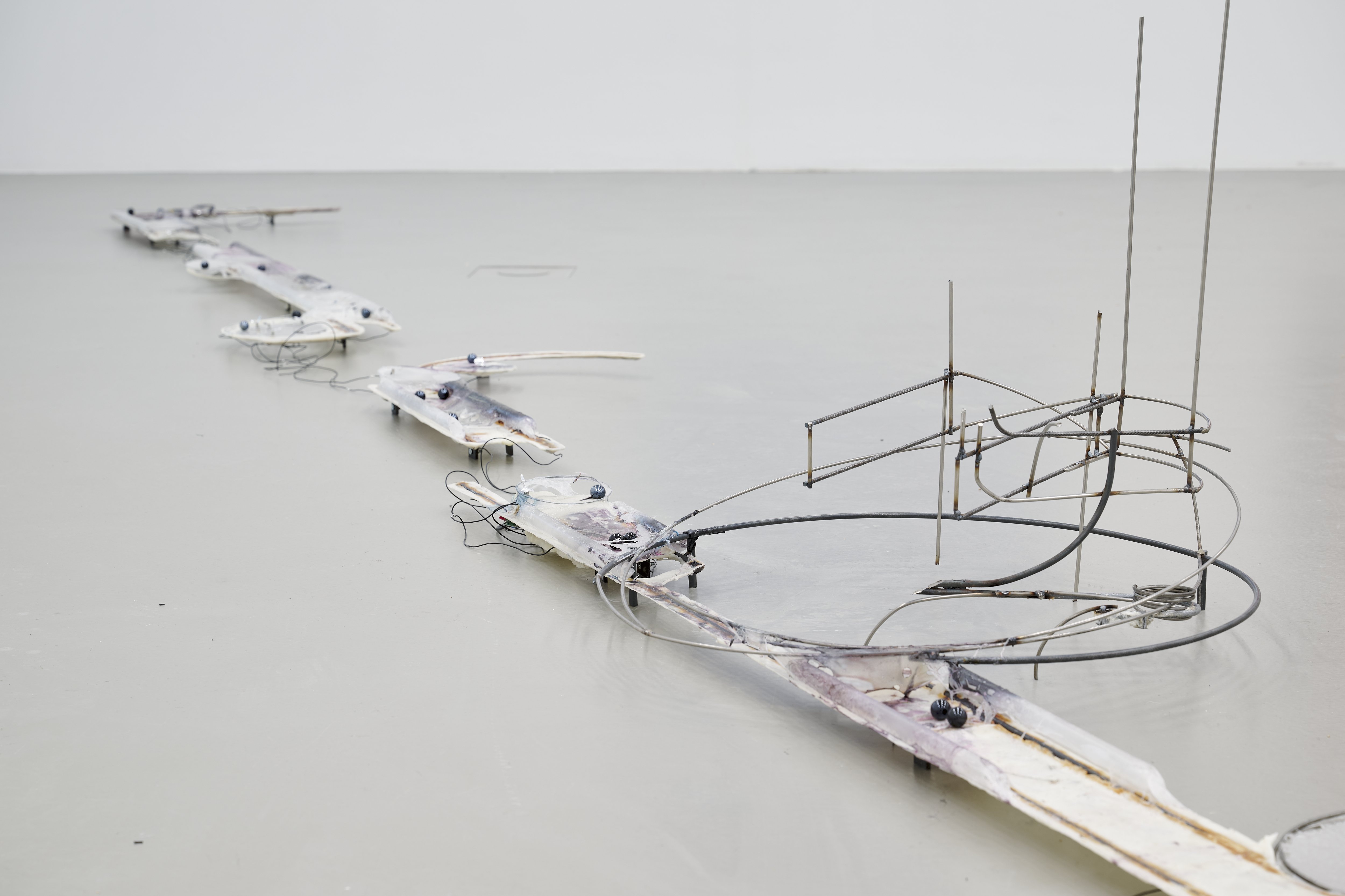 Detailansicht einer langgestreckten Bodenskulptur aus flachen Elementen, Drähten und Metallstrukturen, die sich geschwungen über den Ausstellungsboden erstreckt.