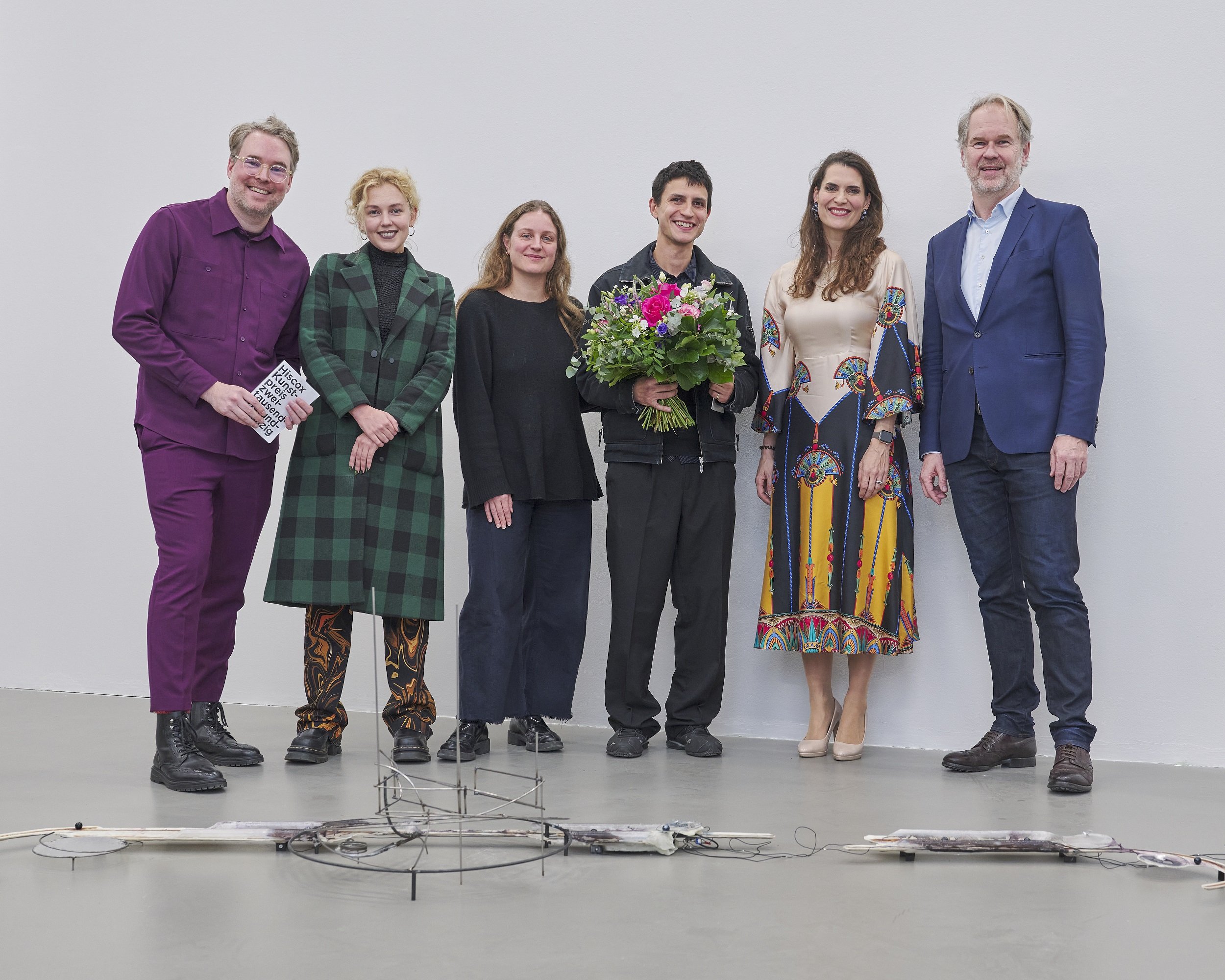 Sechs Personen stehen nebeneinander vor einer weißen Wand; eine Person hält einen Blumenstrauß. Am Boden liegt eine langgestreckte, technische Skulptur mit Draht- und Metallelementen.
