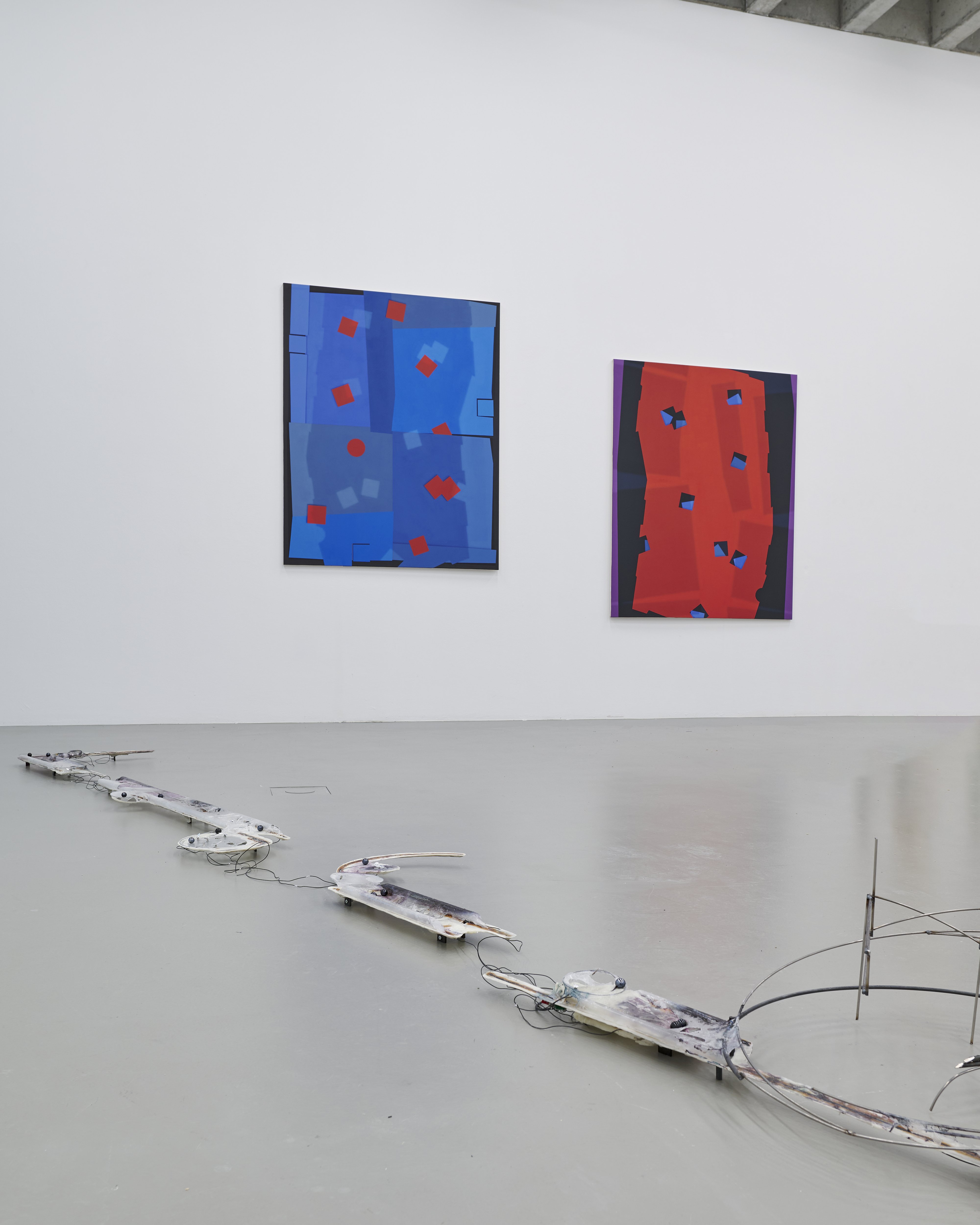 Abstrakte Installation mit einer langgestreckten, technischen Skulptur am Boden, verbunden durch Kabel, vor einer weißen Wand mit zwei farbintensiven abstrakten Gemälden in Blau und Rot.