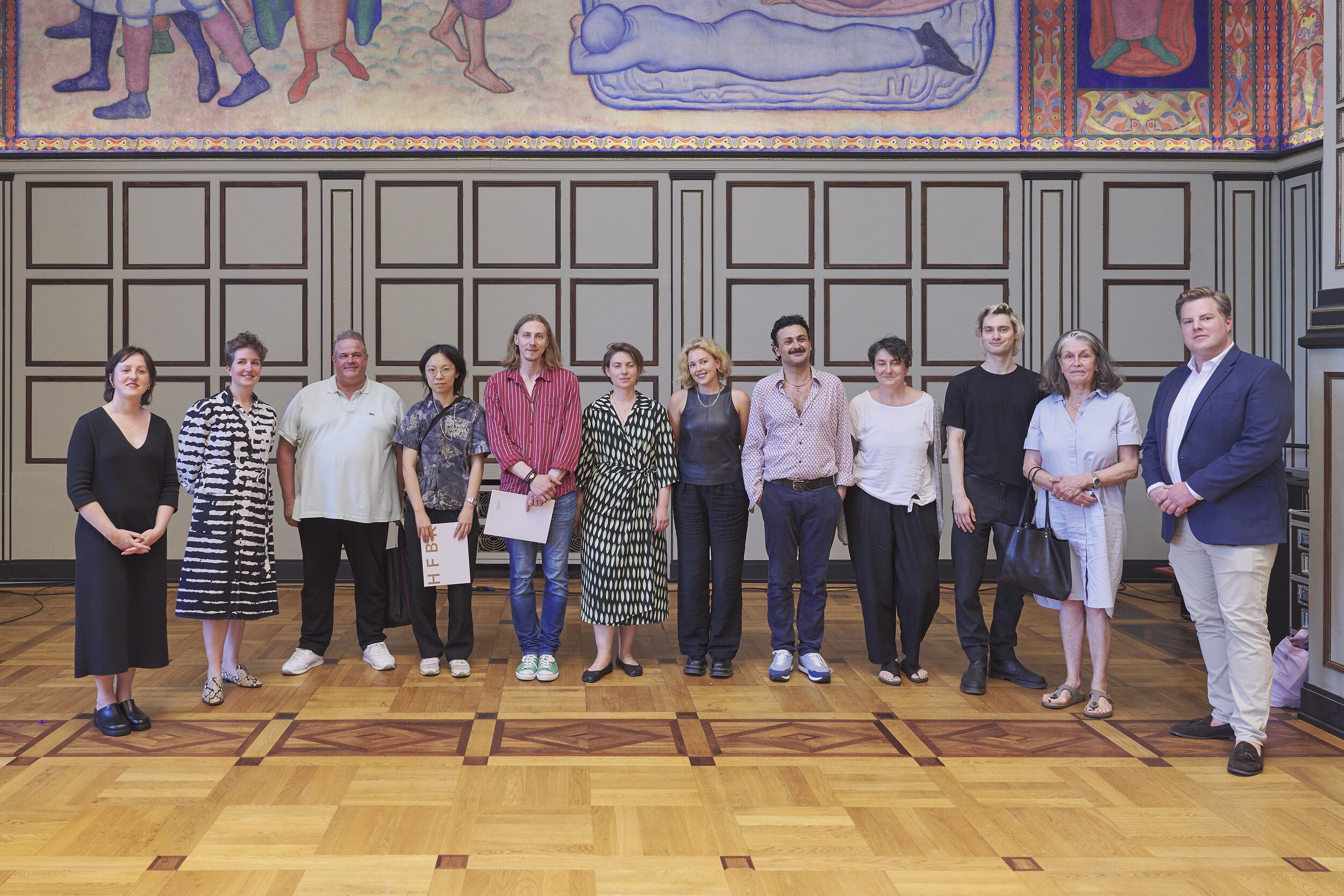 A group of eleven people stands side by side in a historic hall with wooden parquet flooring and paneled walls. A colorful mural is visible above them. The group faces the camera, with some holding bags or documents.