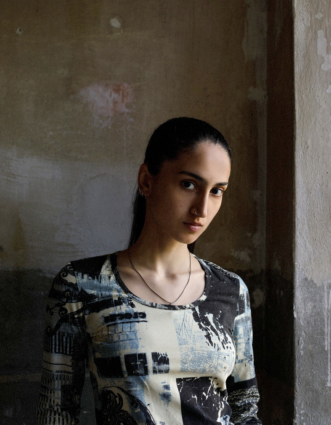 Portrait of a young woman with dark hair pulled back, standing beside a rough concrete wall near a window. Soft side light falls on her face, creating a calm and contemplative mood.
