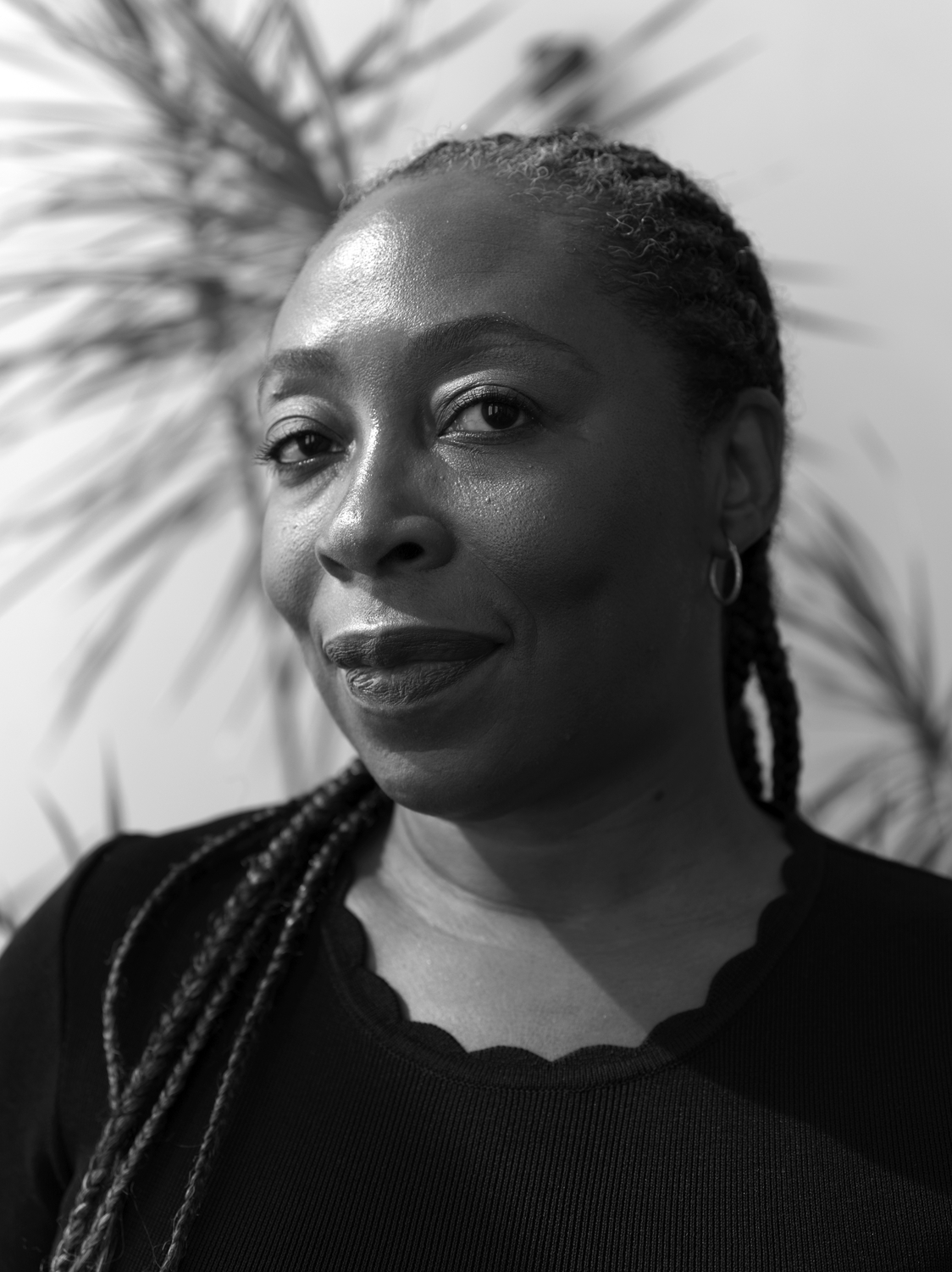 Black-and-white portrait of a woman with braided hair and small earrings. She looks calmly at the camera, with blurred plant leaves visible in the background.