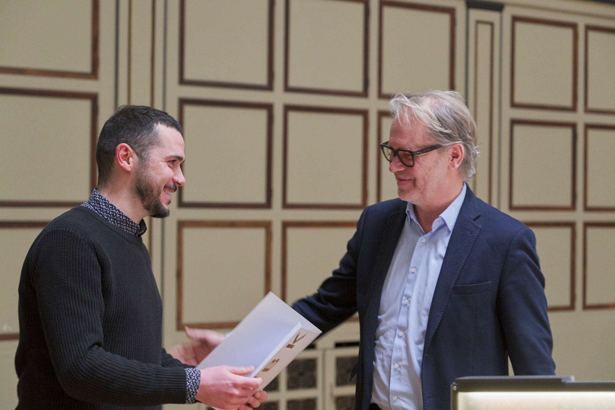 Two people stand facing each other in a hall with wood-paneled walls. One person hands a document or folder to the other, and both are smiling.
