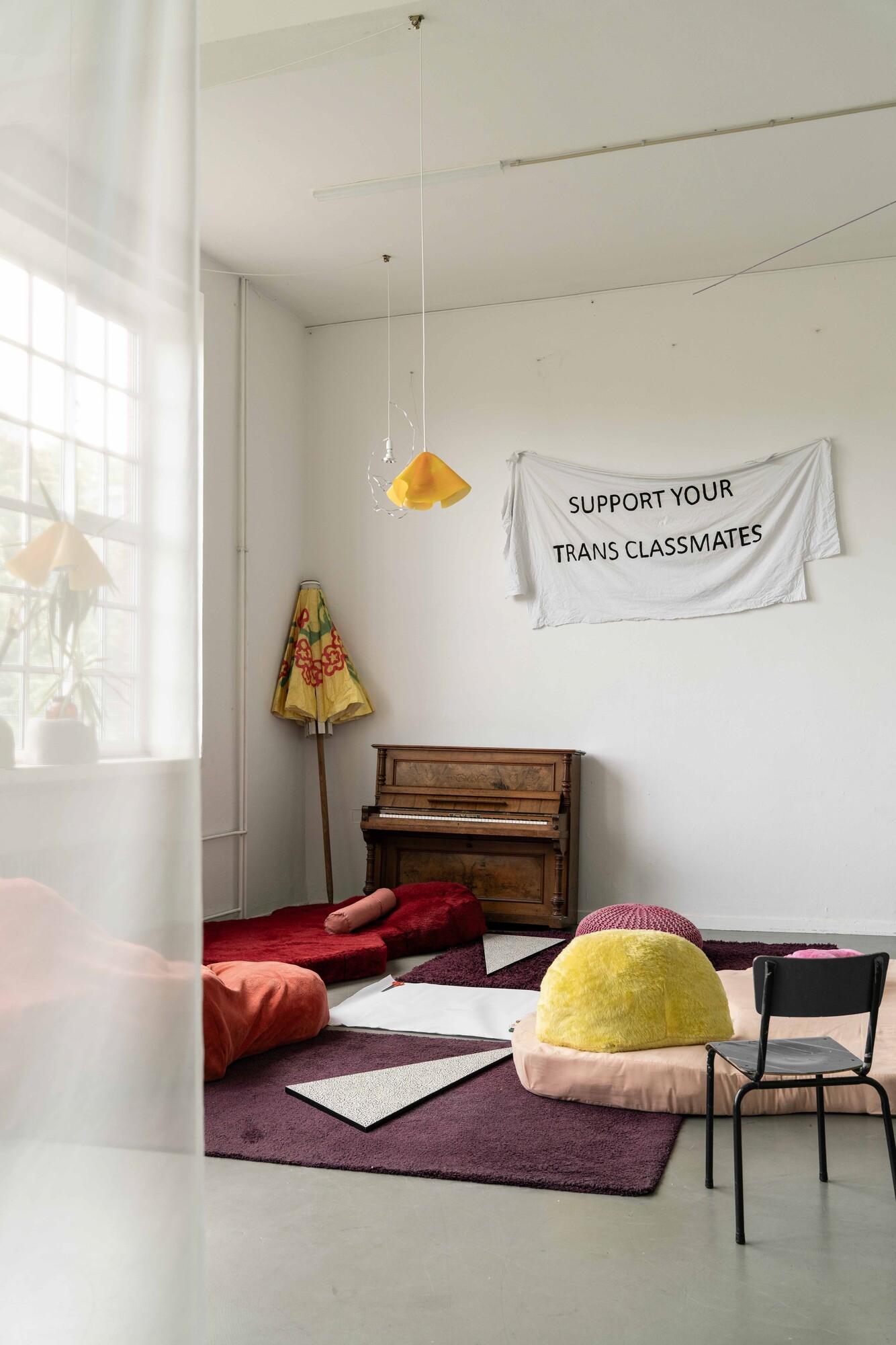 Exhibition space with textile seating and reclining elements placed on rugs, a piano against the wall, and a hanging yellow light fixture; a fabric banner on the wall reads “SUPPORT YOUR TRANS CLASSMATES”.