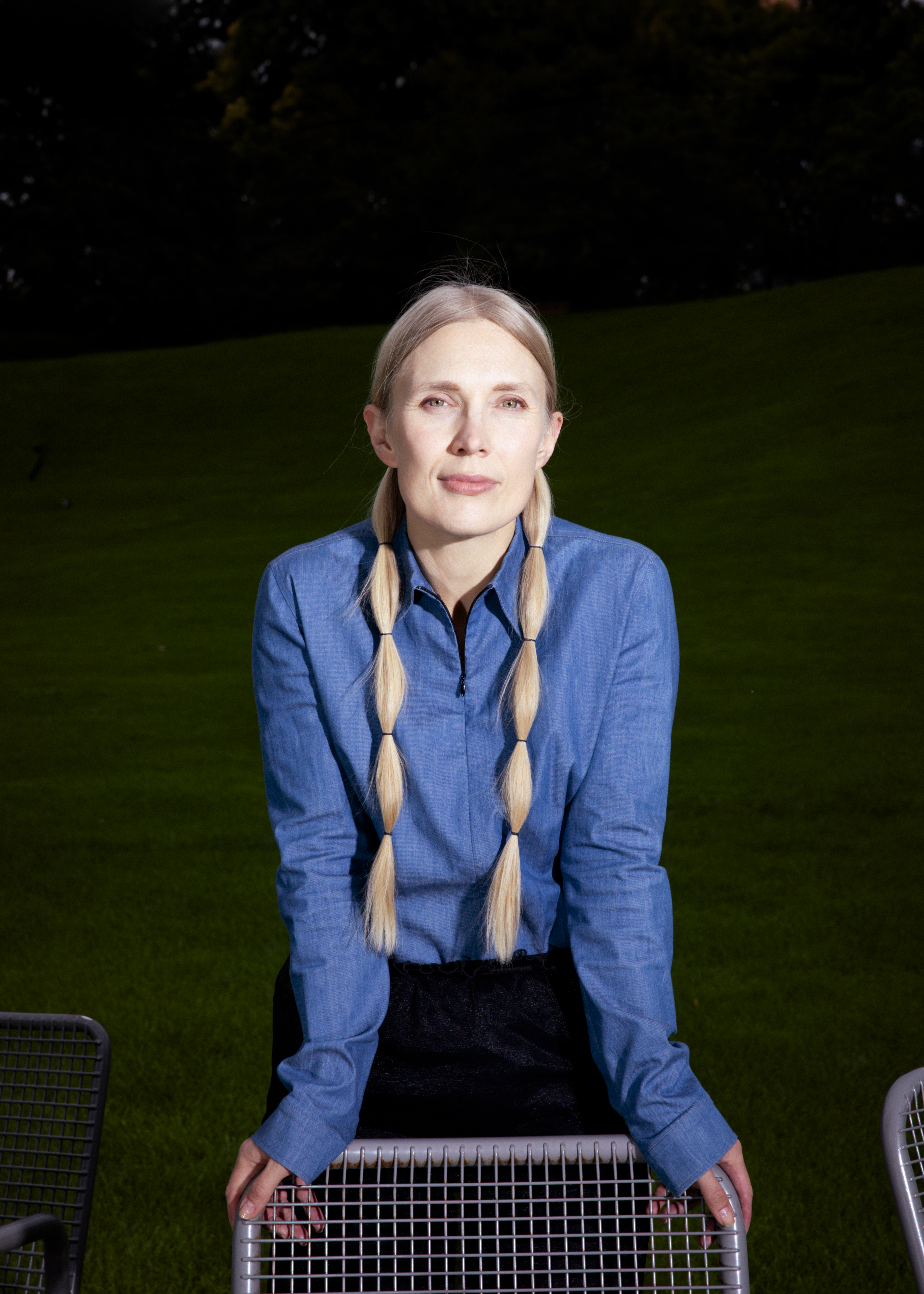 Eine Frau mit langen blonden Zöpfen und einem blauen Hemd stützt sich mit beiden Händen auf die Metalllehne eines Stuhls.