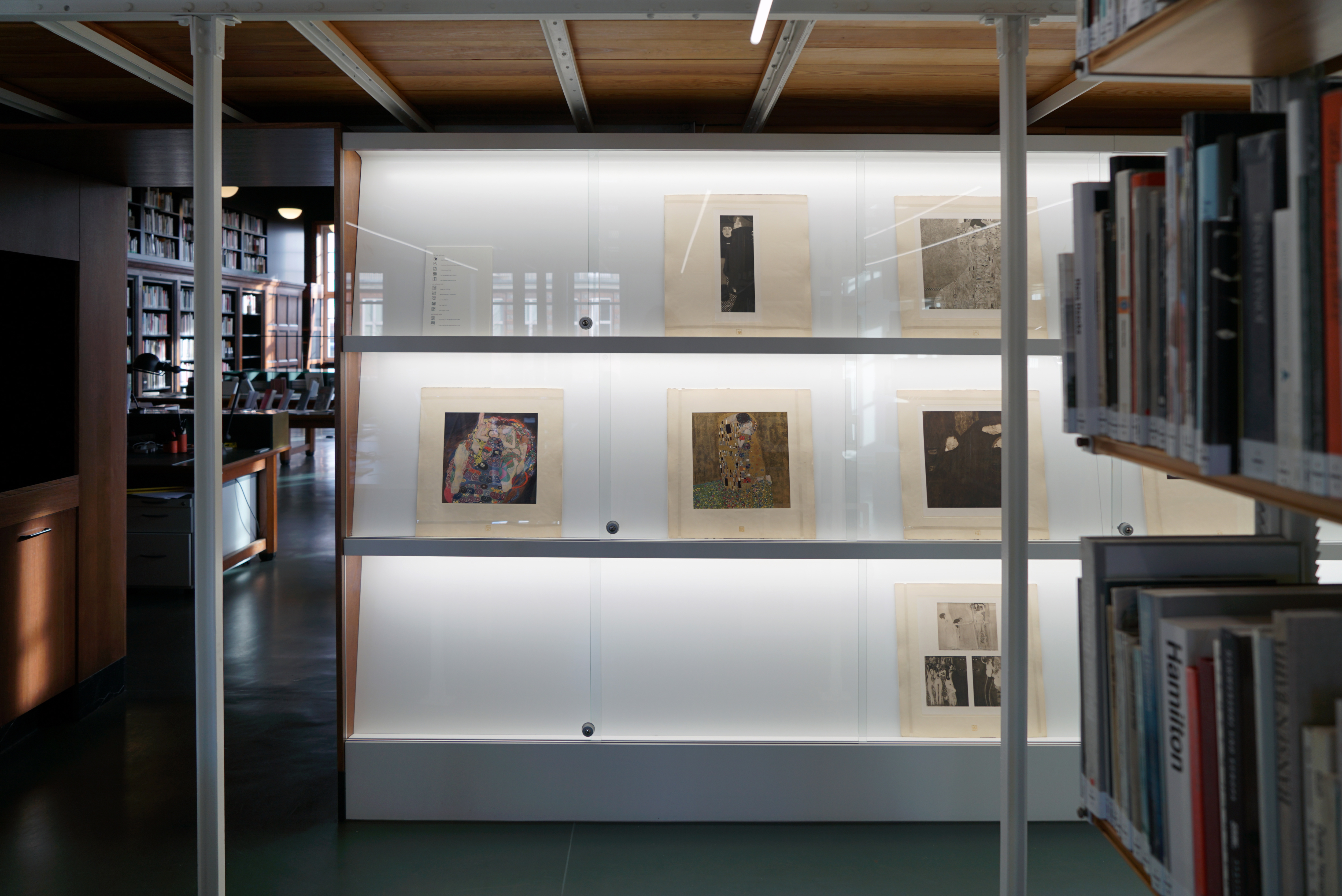 Display case with framed artworks in a library space. Bookshelves are visible in the foreground, and workstations and shelves are in the background.