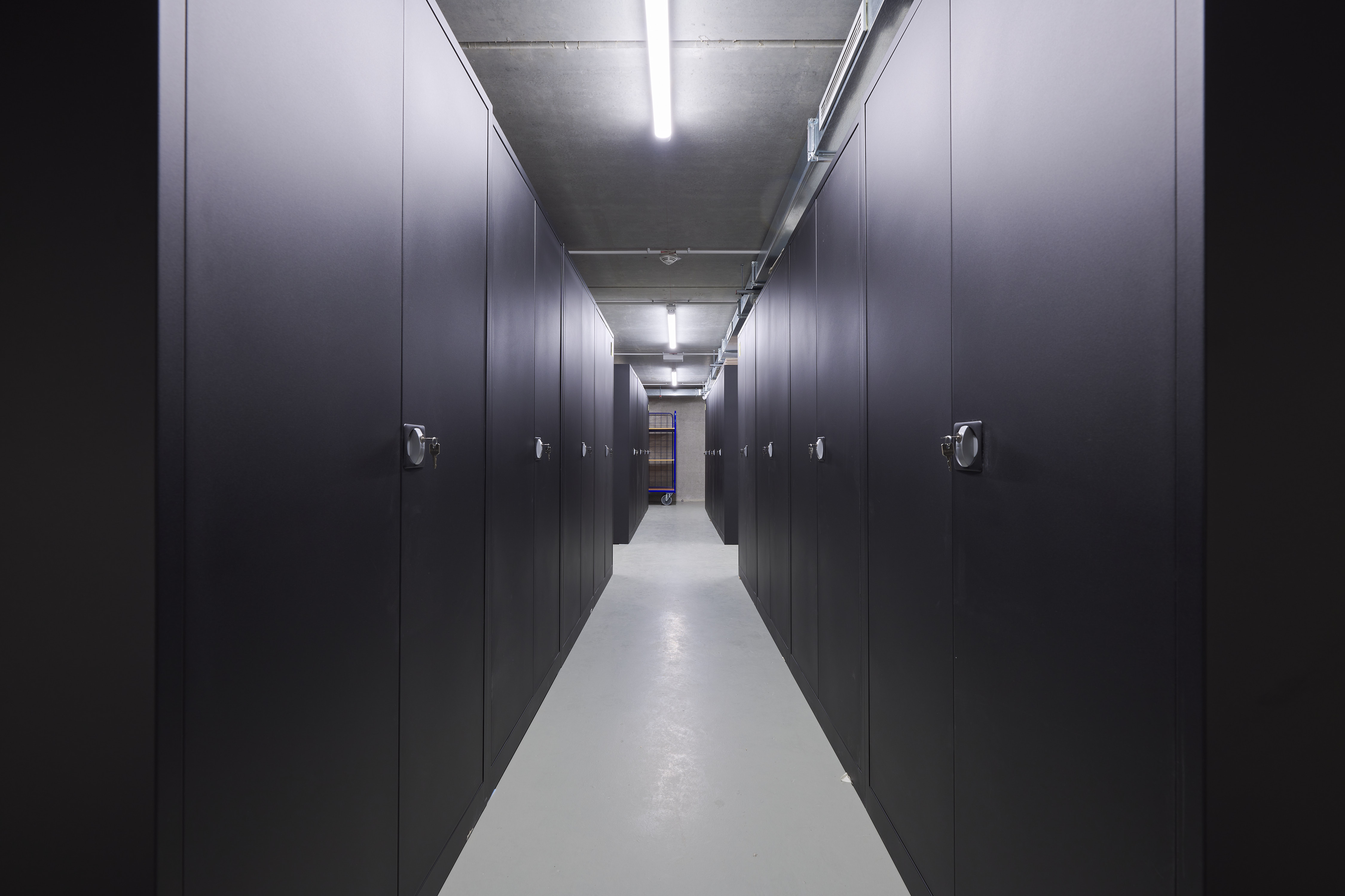 A narrow corridor in an archive or storage room, lined with tall dark-grey metal cabinets equipped with turning handles. The concrete ceiling features exposed cables and a long fluorescent light. At the end of the corridor, a cart stands in front of a mesh door.
