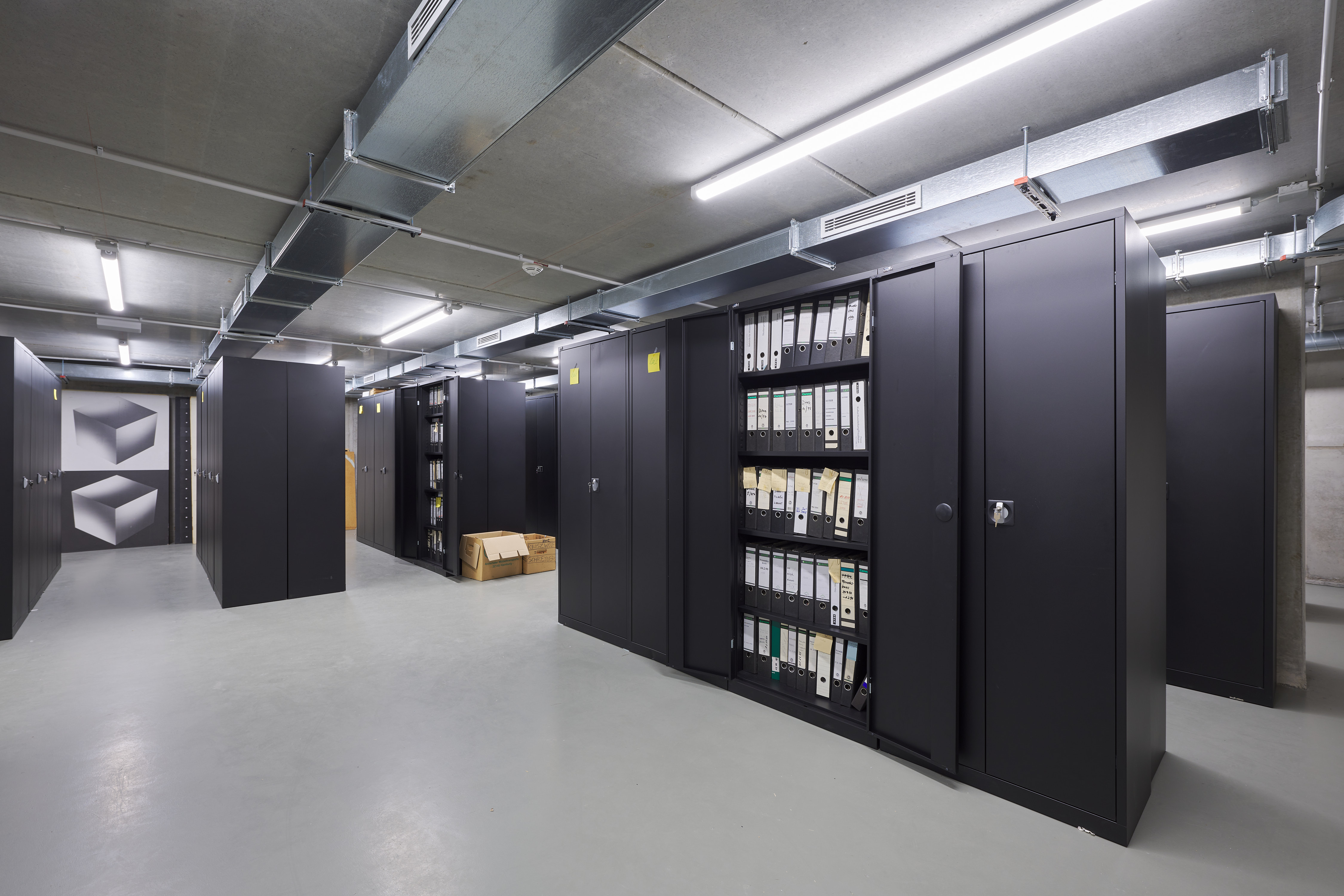 Large, well-lit archive room with dark metal cabinets and rows of file folders. Ventilation pipes run along the ceiling.