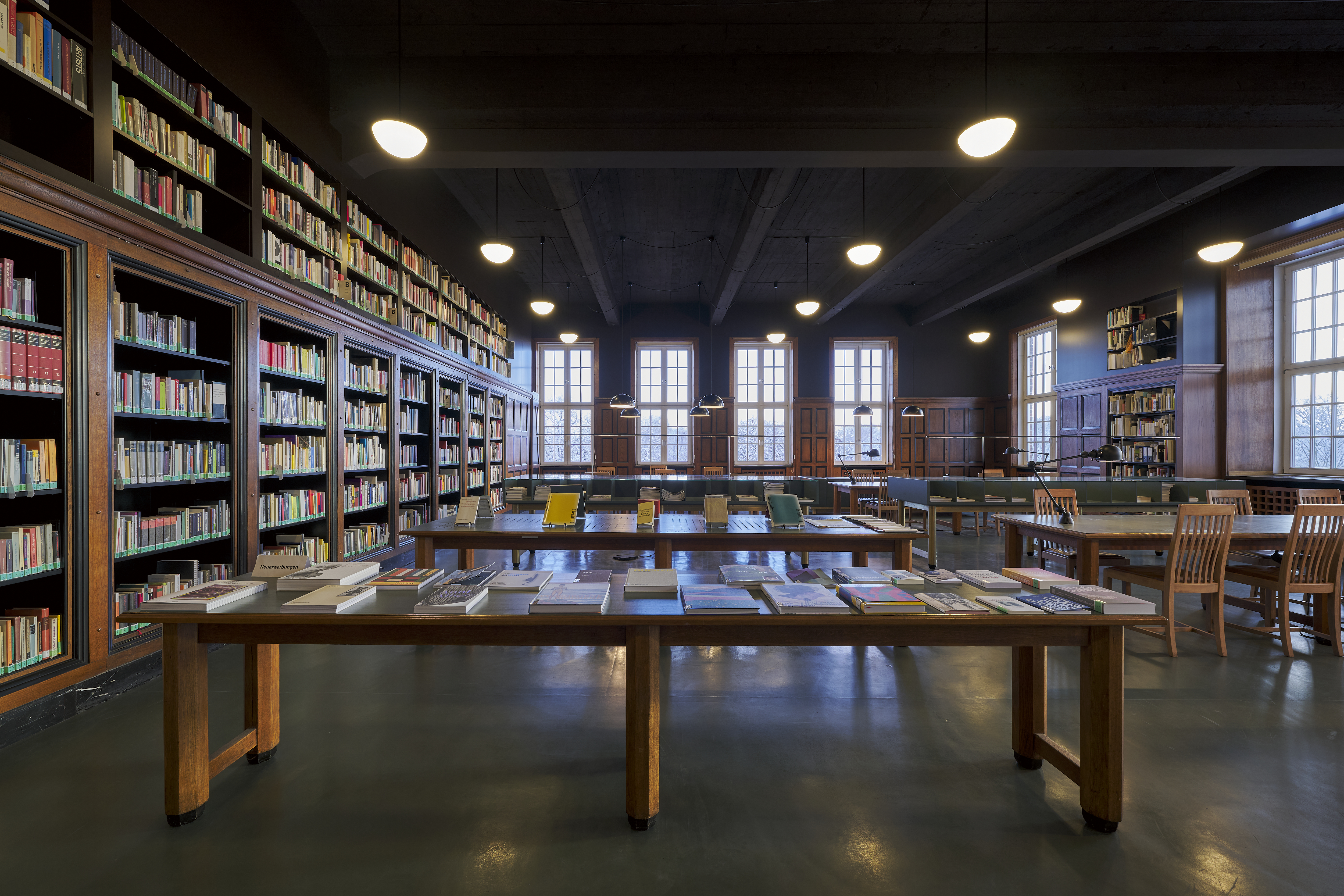 Sideways ceiling-high bookshelf, tables with books laid out in the center, tables with chairs on the right-hand side