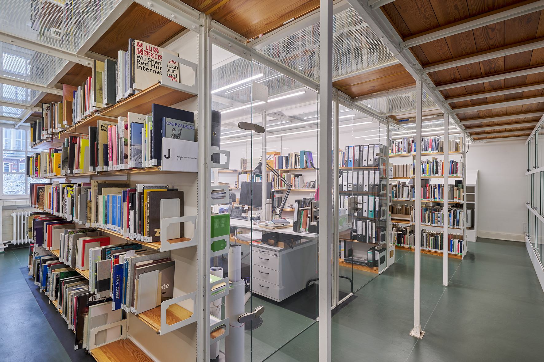 Innenraum einer modernen Bibliothek oder eines Archivs mit hohen Holzregalen voller Bücher, verglasten Büroabteilen und einem Arbeitsplatz mit Schreibtisch im Inneren. Der Raum hat eine weiße Metallkonstruktion, Holzdeckenpaneele und einen grünen Boden.