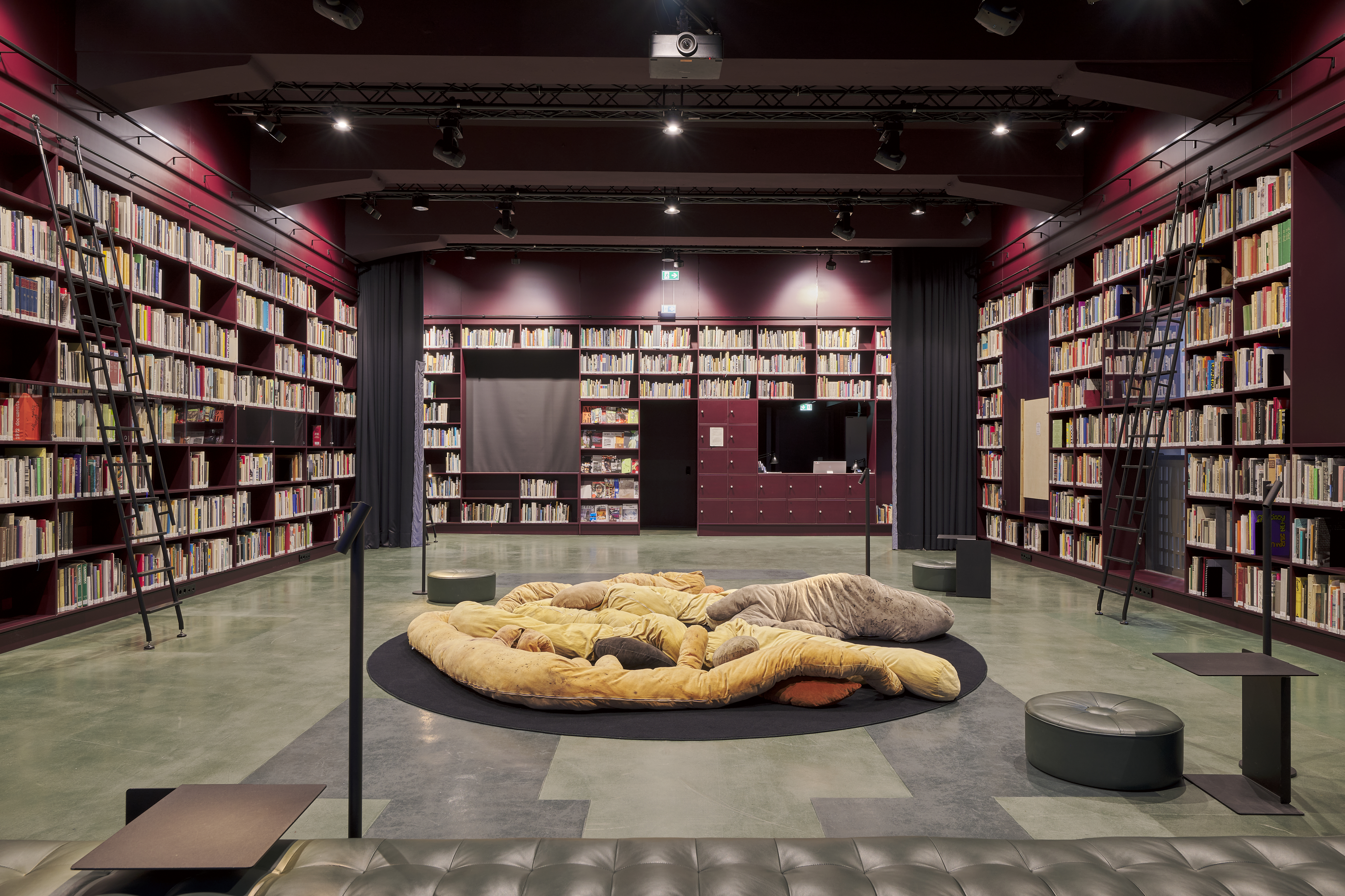 Room with many books on the walls, reception desk in the rear right corner, and a lawn area with a cushion object in the center