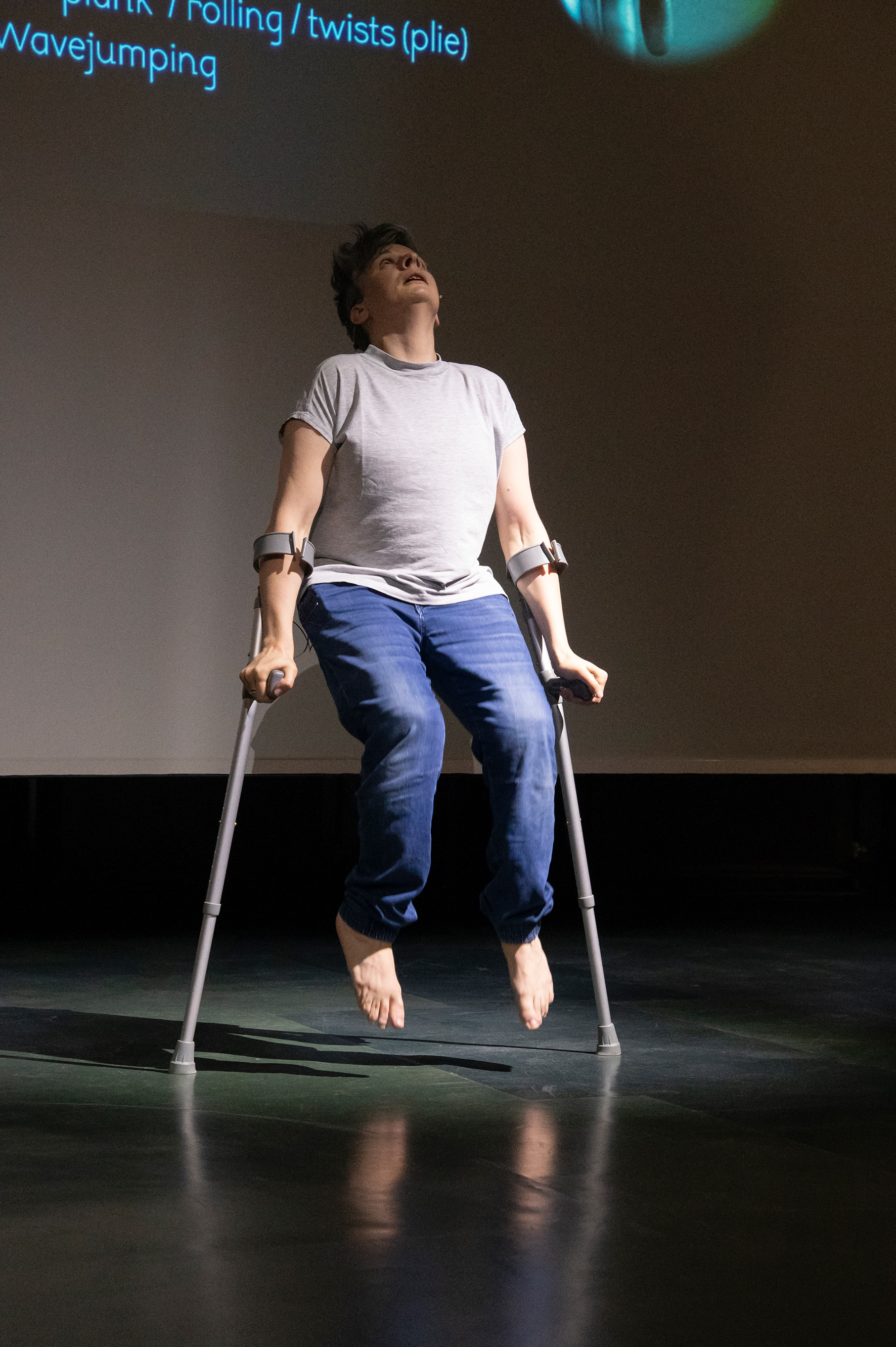 A person stands barefoot on a stage, supported by two forearm crutches, tilting their head upward. Text fragments are projected onto the screen in the background.