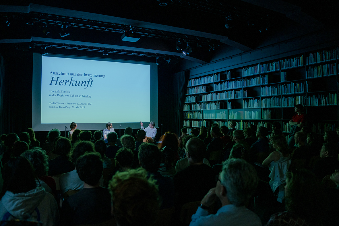An audience sits in a dimly lit room facing a large projection screen displaying the title “Excerpt from the stage production Herkunft.” Three people are seated at the front in discussion, with floor-to-ceiling bookshelves visible in the background.
