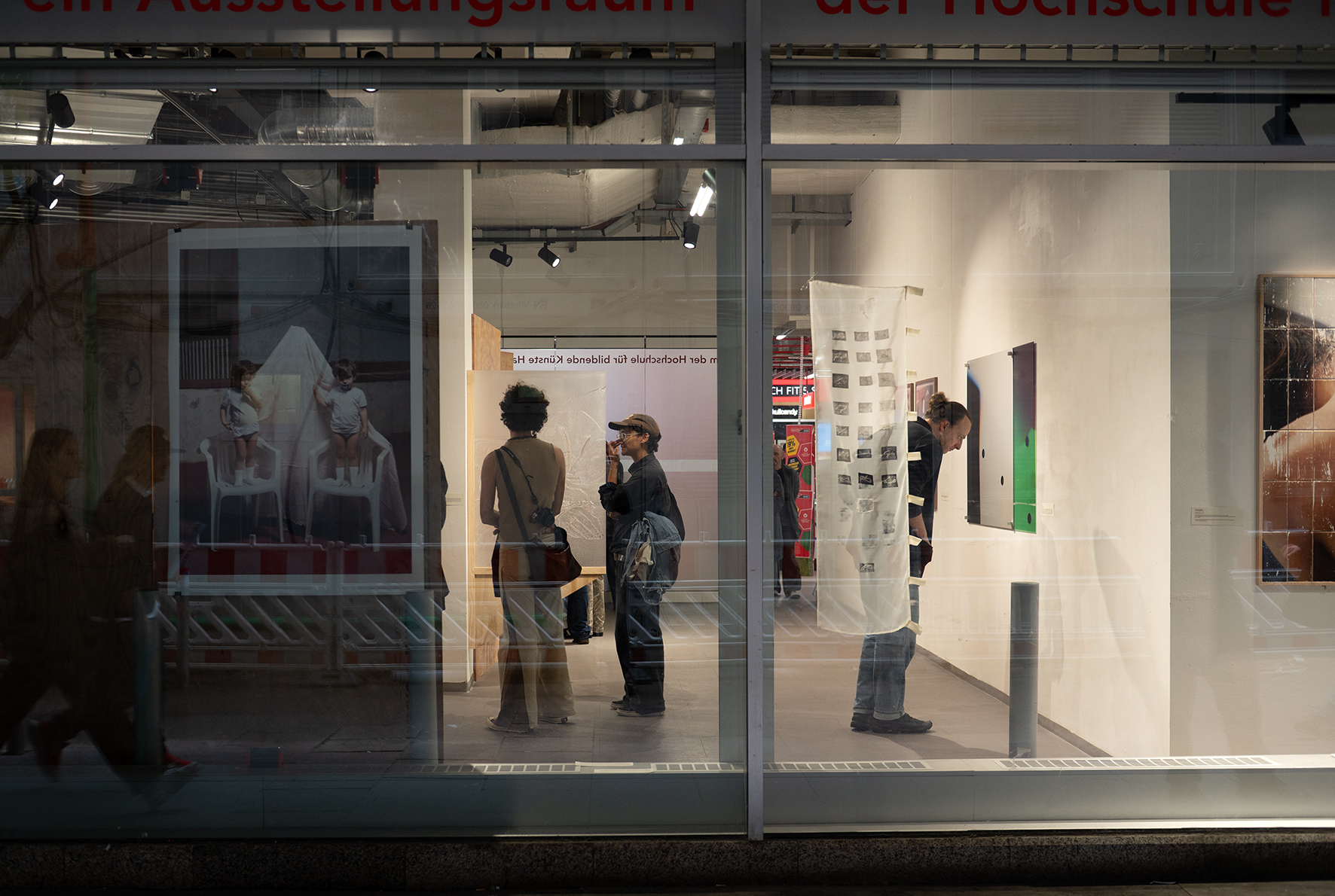 Blick durch die Scheibe ins Innere des Ausstellungsraumes, in dem Menschen stehen und sich Fotografien anschauen.