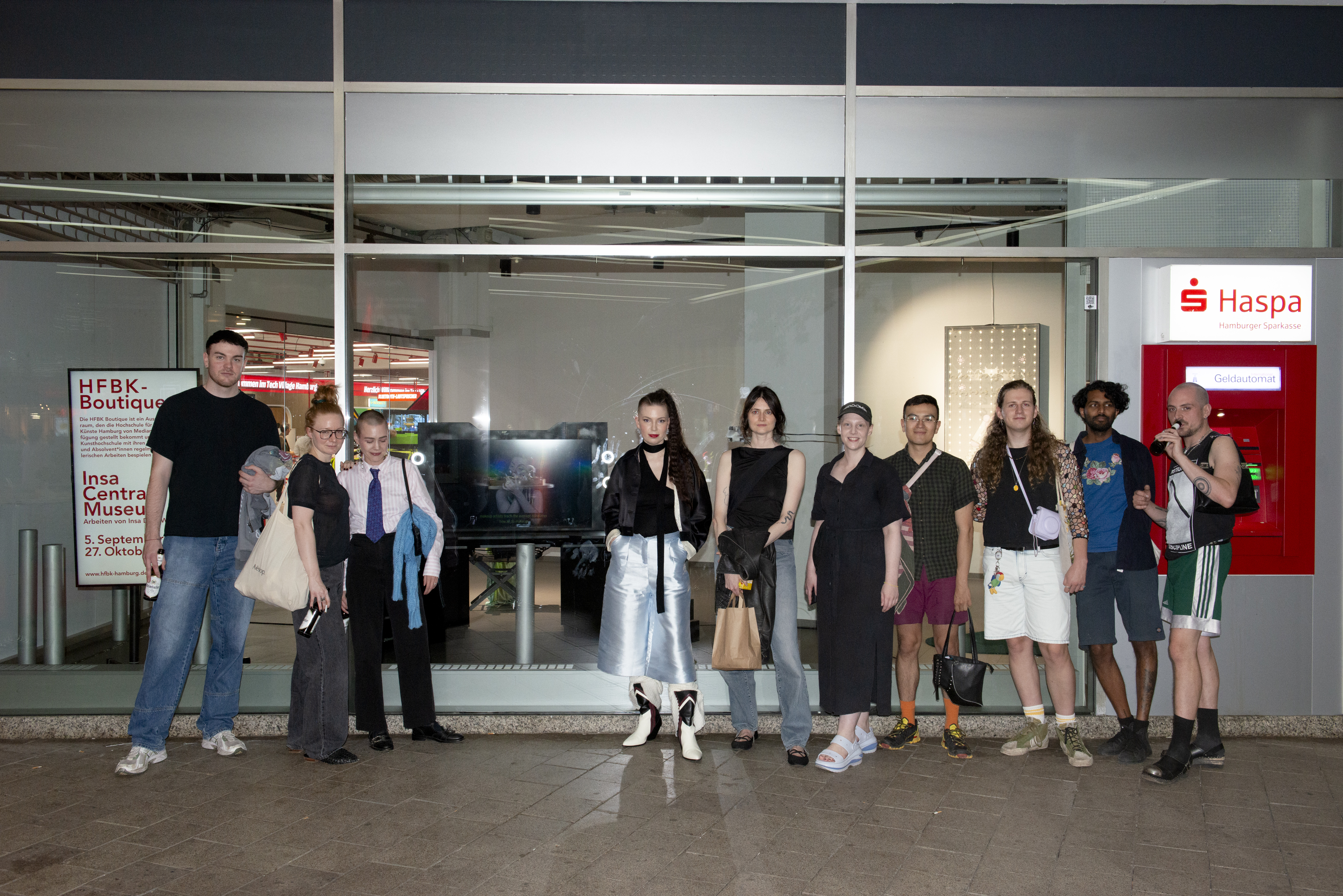 A group of young people standing in front of a shop window next to a savings bank ATM