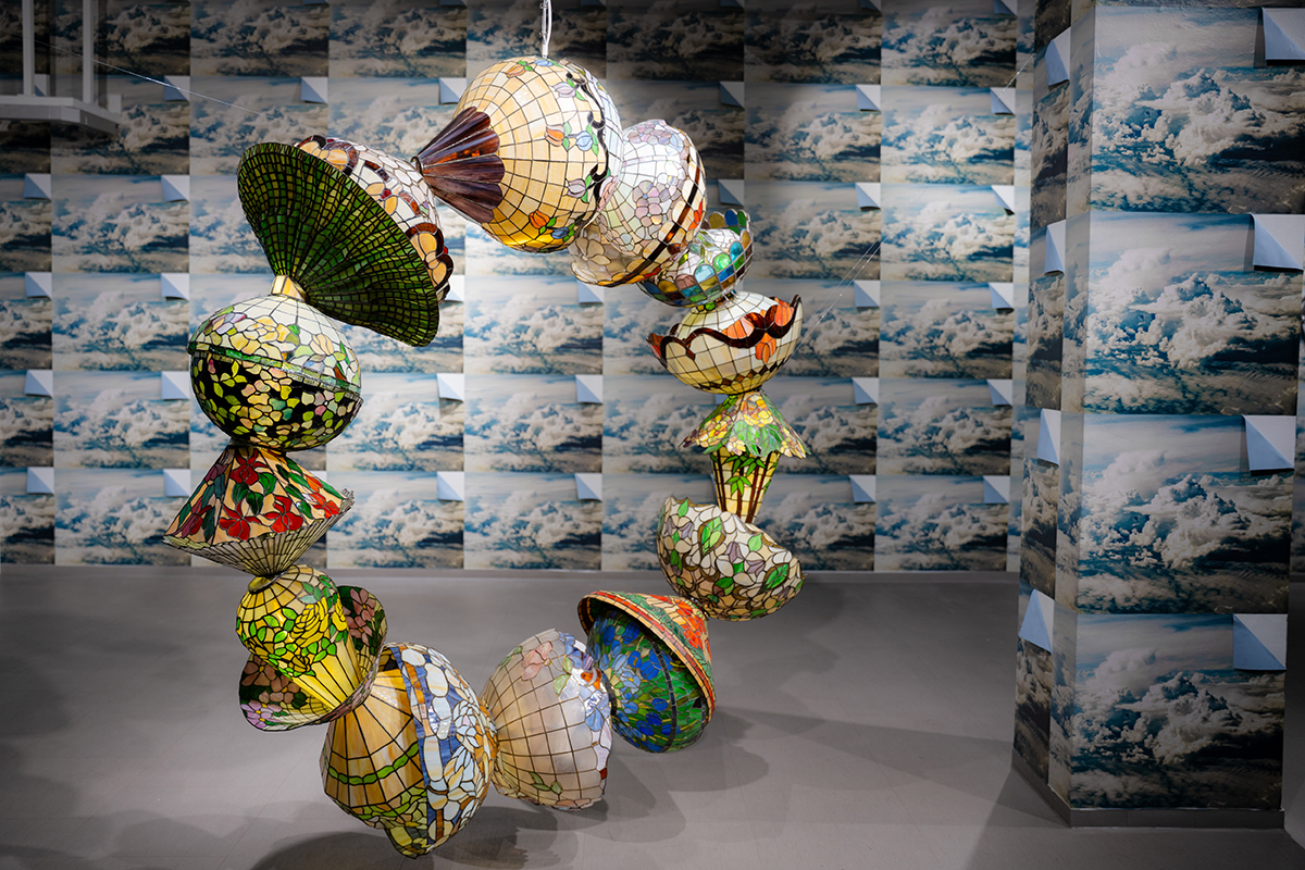 A circular sculpture made of lampshades hangs in a room, with photo wallpaper on the wall.