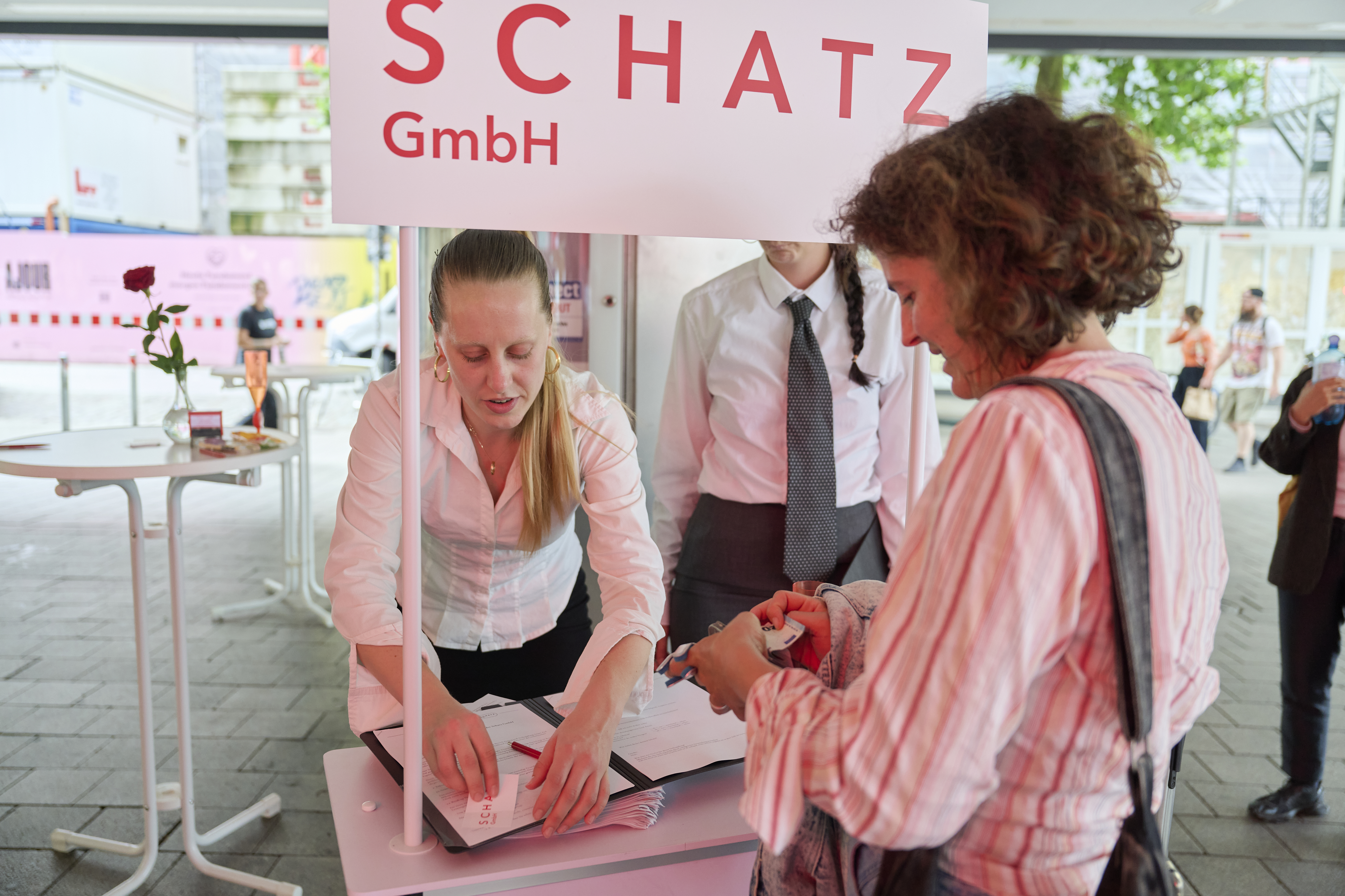 Two women are standing at an information stand with a sign that reads “Schatz GmbH,” an artistic dating agency.