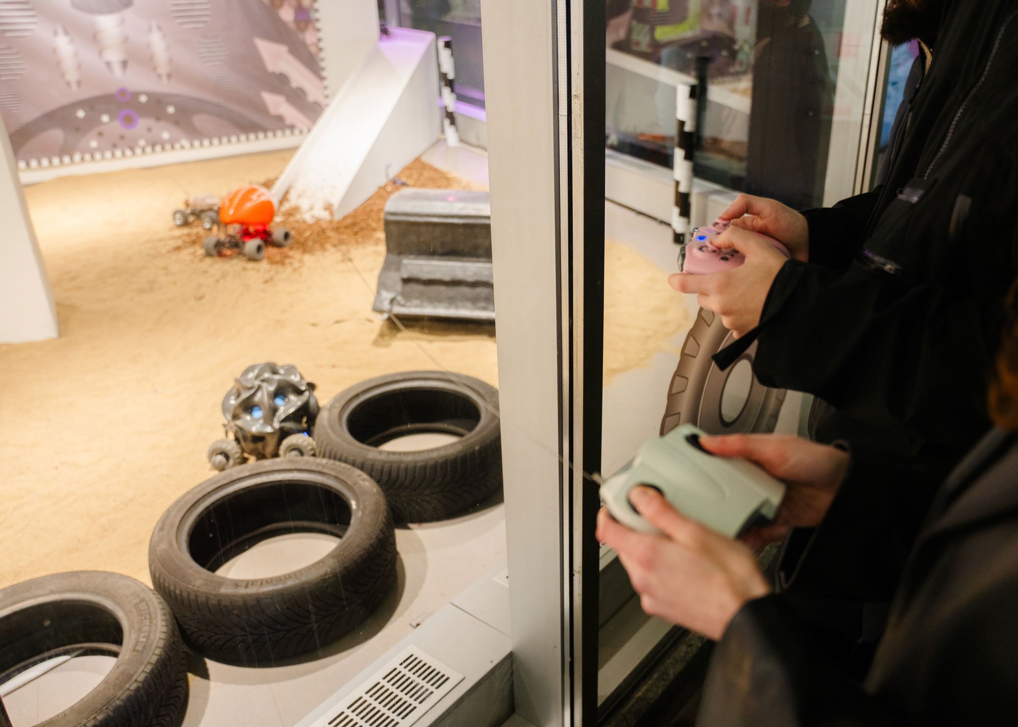 Two people stand in front of a glass enclosure, using handheld controllers to steer small vehicles inside a sandy arena. Several car tires are arranged as obstacles, and small remote-controlled cars—one black and one orange—drive across the sand. The scene appears to be part of an interactive exhibition or play installation.
