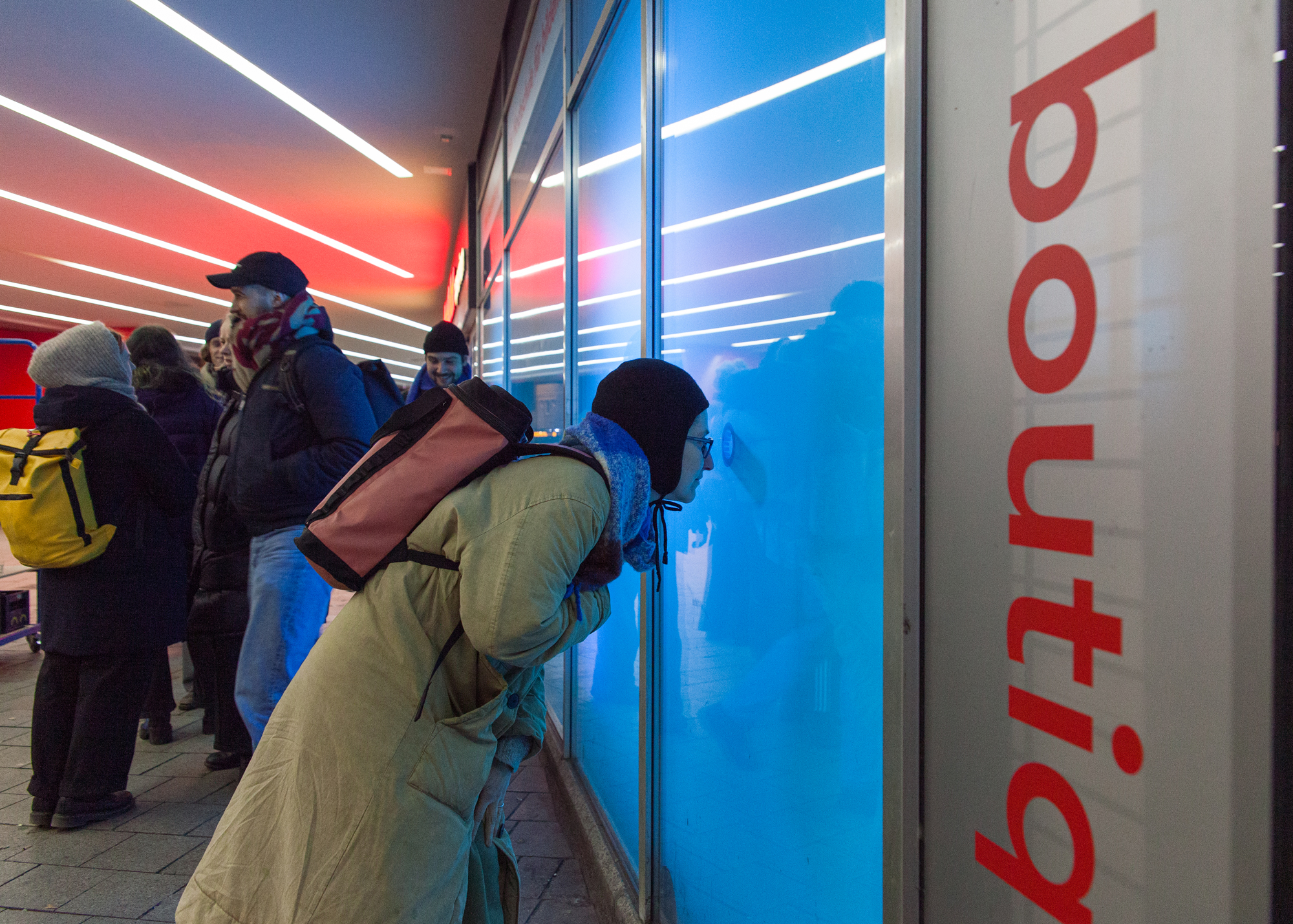 Several people stand outside a glass façade at night illuminated by blue light. One person wearing a backpack and winter clothing leans forward to peer curiously through the window. Neon strip lights cast red and white light across the entrance area, and the word “boutiq” appears vertically on the right.