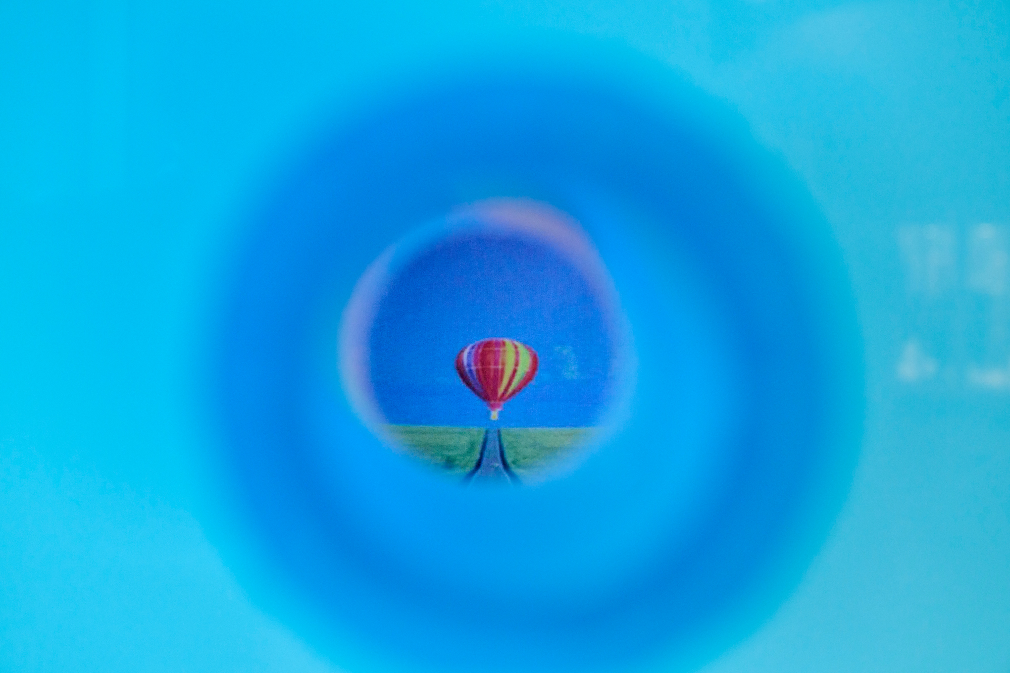 Through a circular, blue-blurred opening, a small image appears at the center: a red-and-yellow striped hot air balloon floating above a flat green landscape under a blue sky. The edges of the frame are softly blurred in turquoise tones.