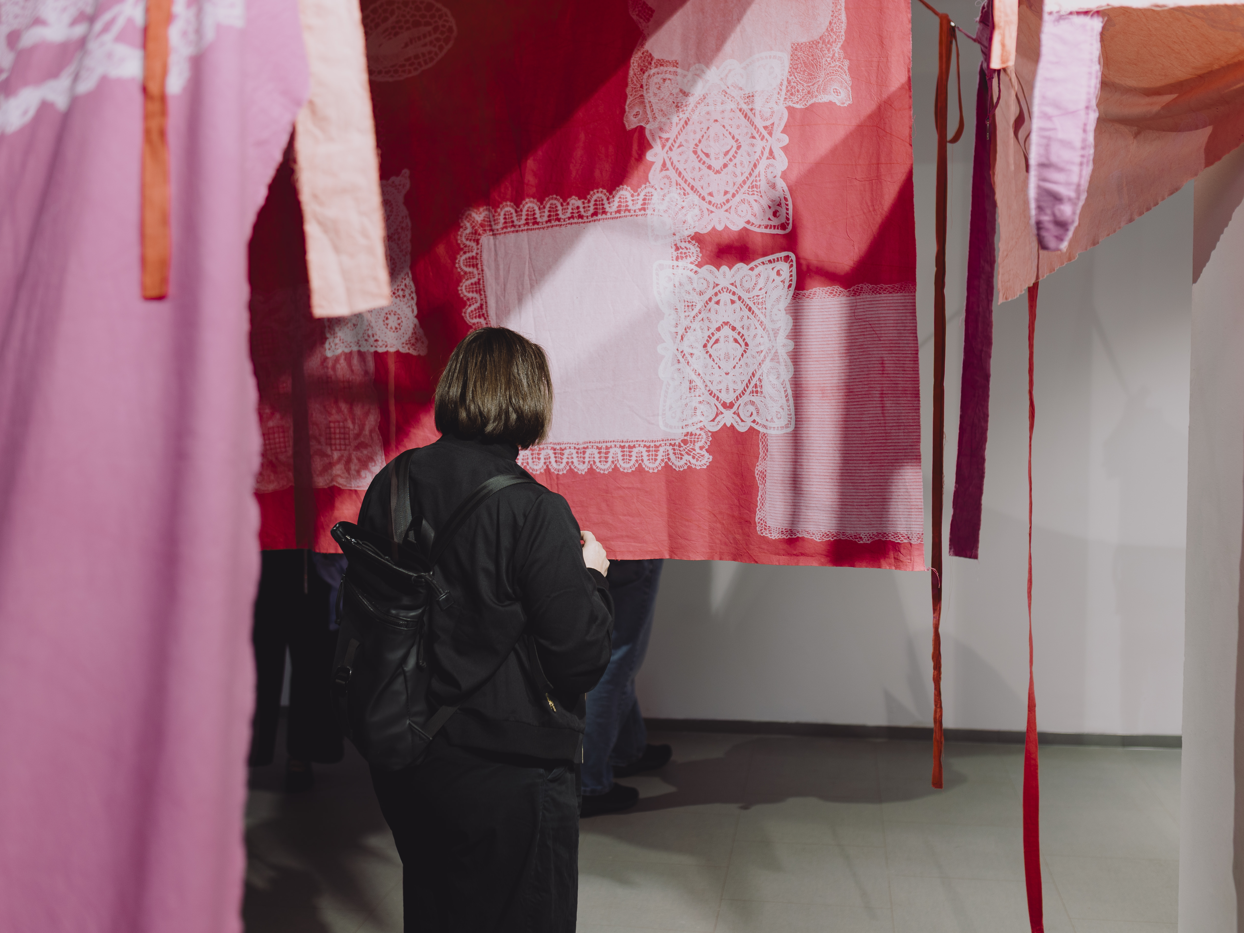 Person with black backpack stands in a room with hanging red and pink fabrics featuring lace patterns