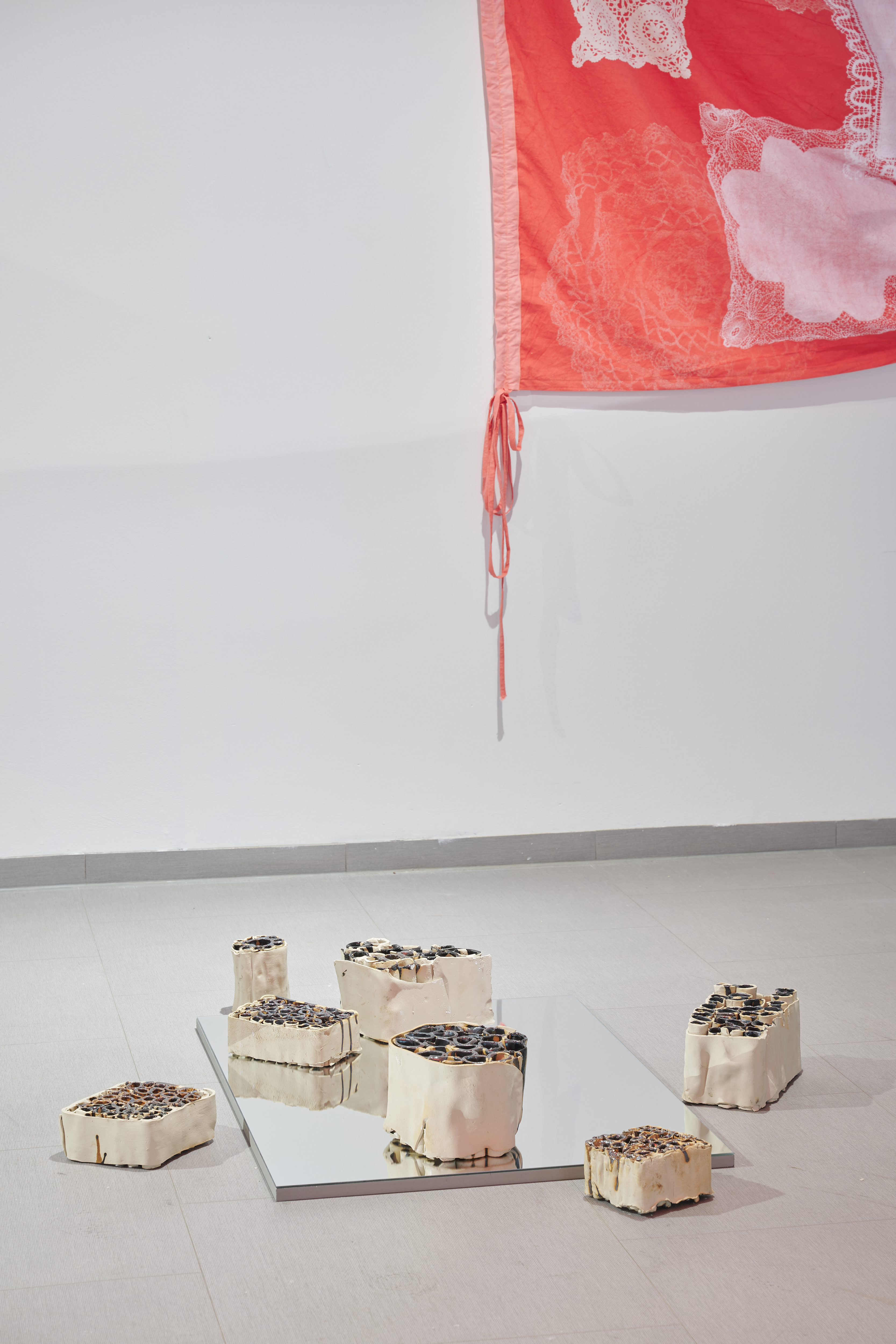 Several porous, irregularly shaped containers made of light material placed on the floor, some on a rectangular mirror, with a red-and-white cloth hanging on the wall in the background