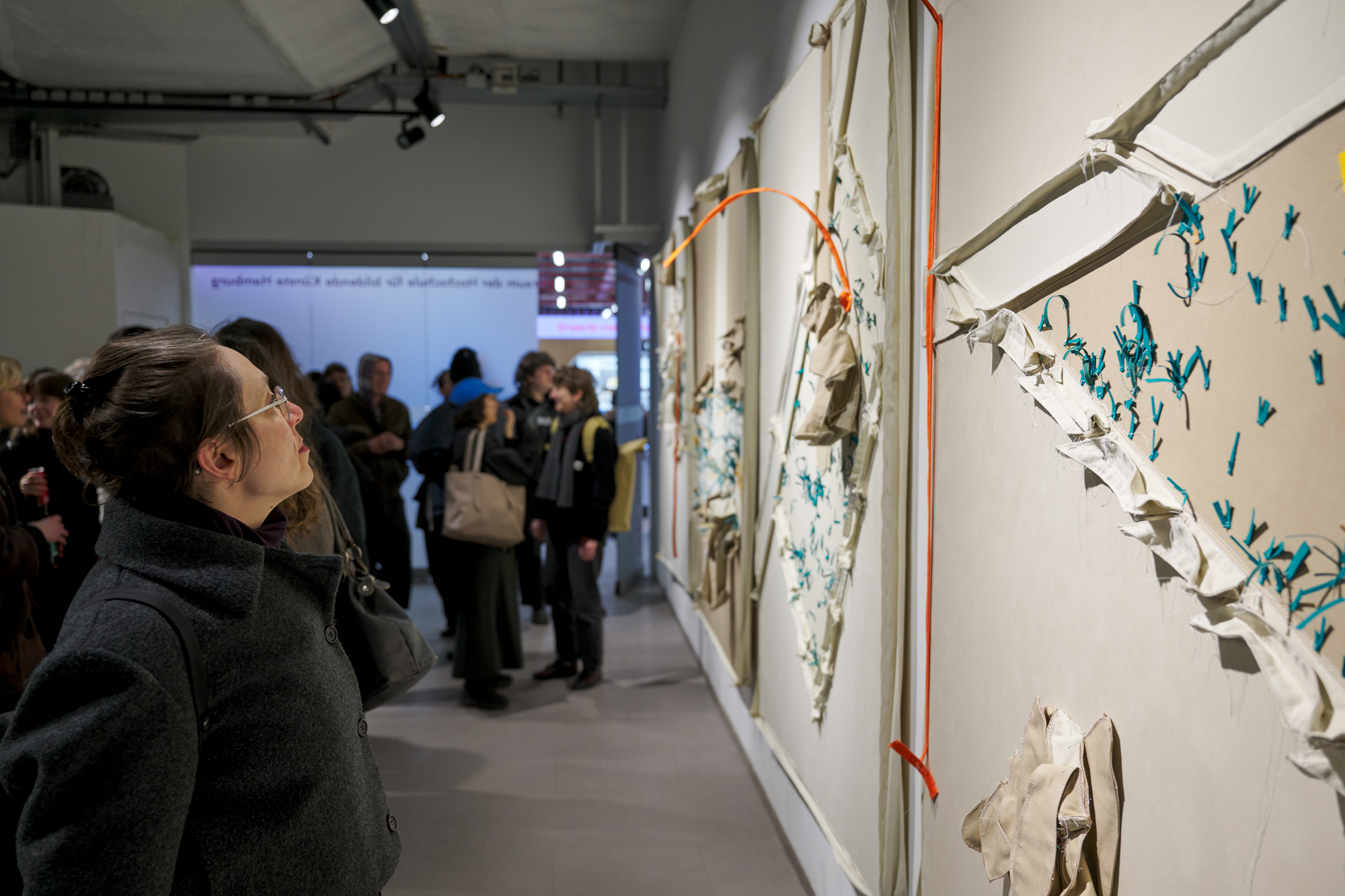 A woman closely examines a large abstract artwork on display in a gallery, while other people gather and converse in the background.