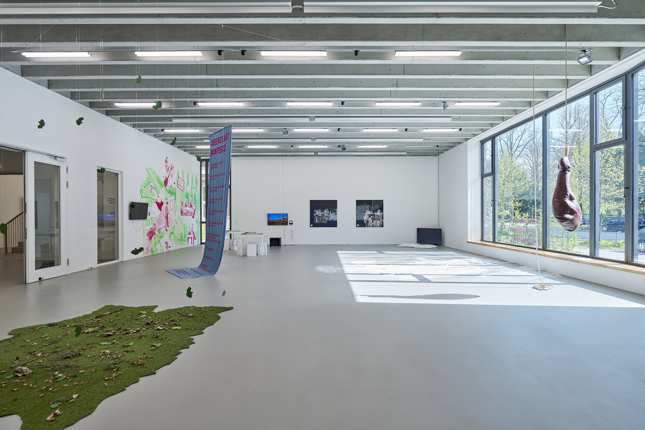 A bright exhibition space with large windows on the right and various installations, including a suspended fabric banner with text, wall works, video monitors, and a patch of artificial grass with leaves on the floor. A sculptural object hangs in front of the window on the right.