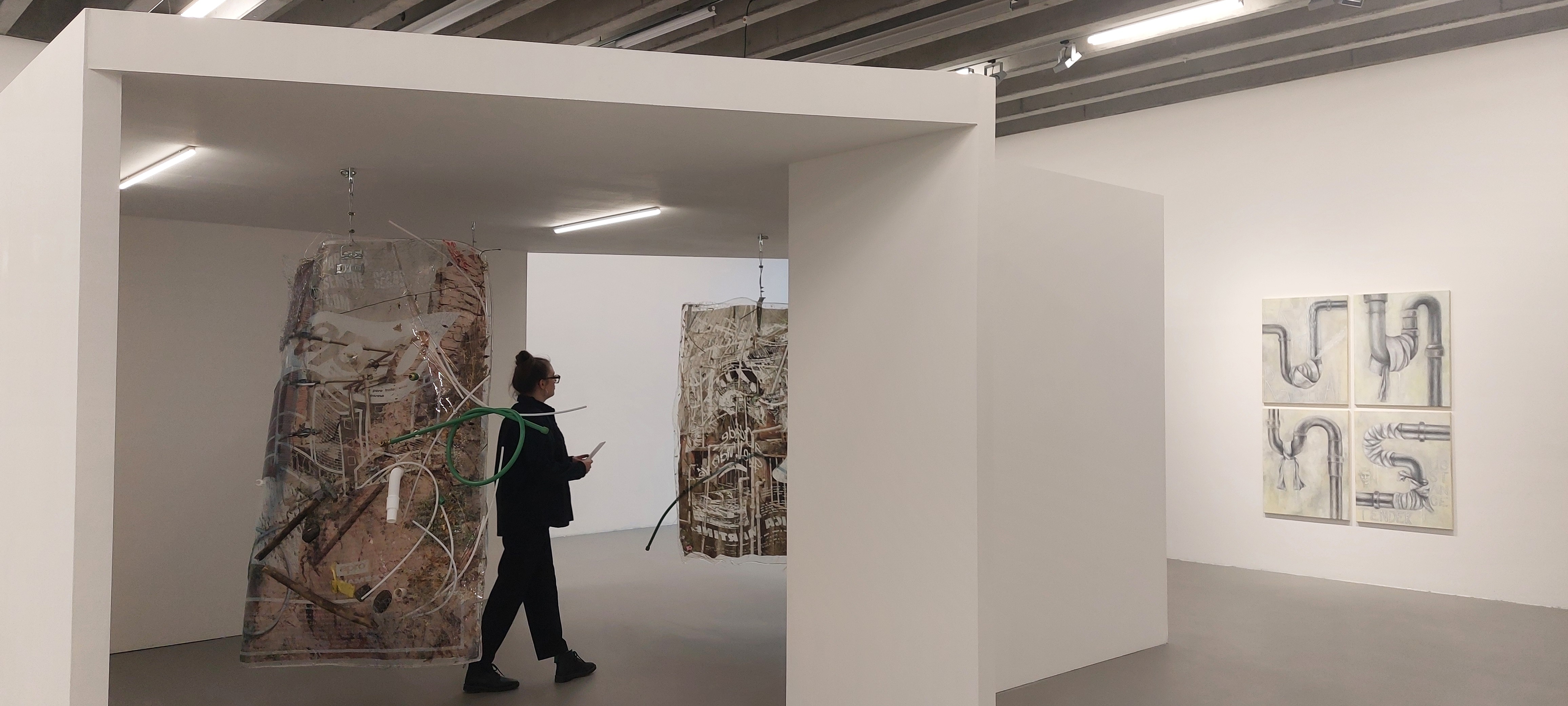 Eine Person läuft zwischen hängenden Kunstwerken hindurch, an der rechten Wand sind Bilder mit gemalten Rohren zu erkennen.