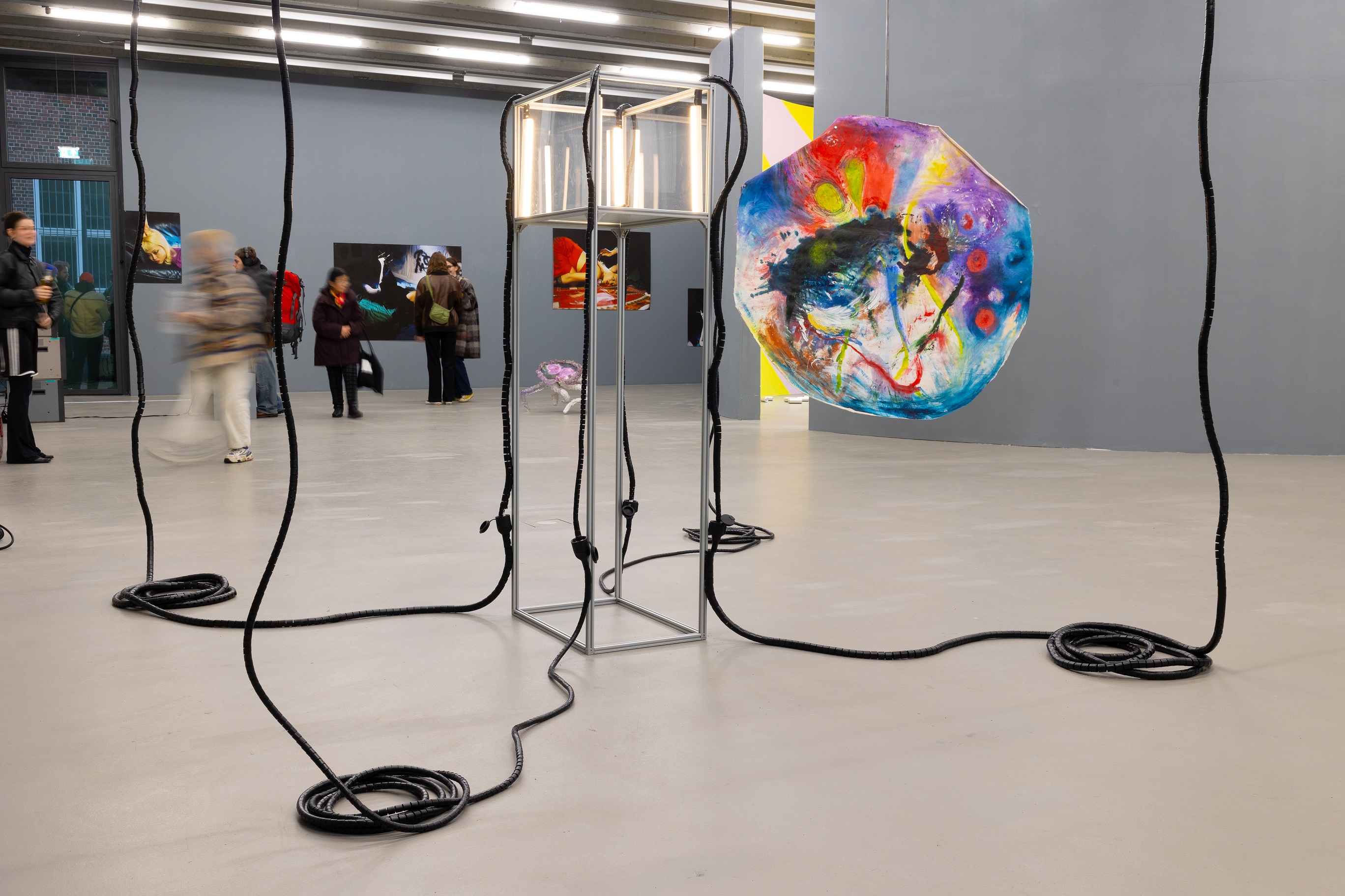 A large exhibition space featuring an installation of black cables hanging from the ceiling and coiled on the floor. In the center stands a glowing cube-like frame, and to the right hangs a large, vividly colored circular painting. In the background, visitors view additional artworks on the walls.