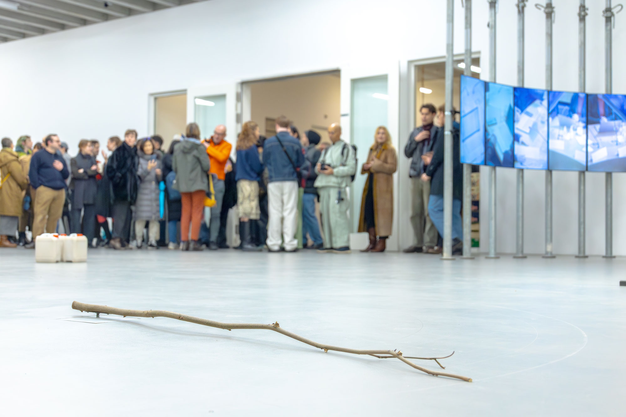 In a bright exhibition space, a single branch lies on the floor in the foreground. In the background, visitors stand together in groups, and several monitors displaying video works are visible on the right.