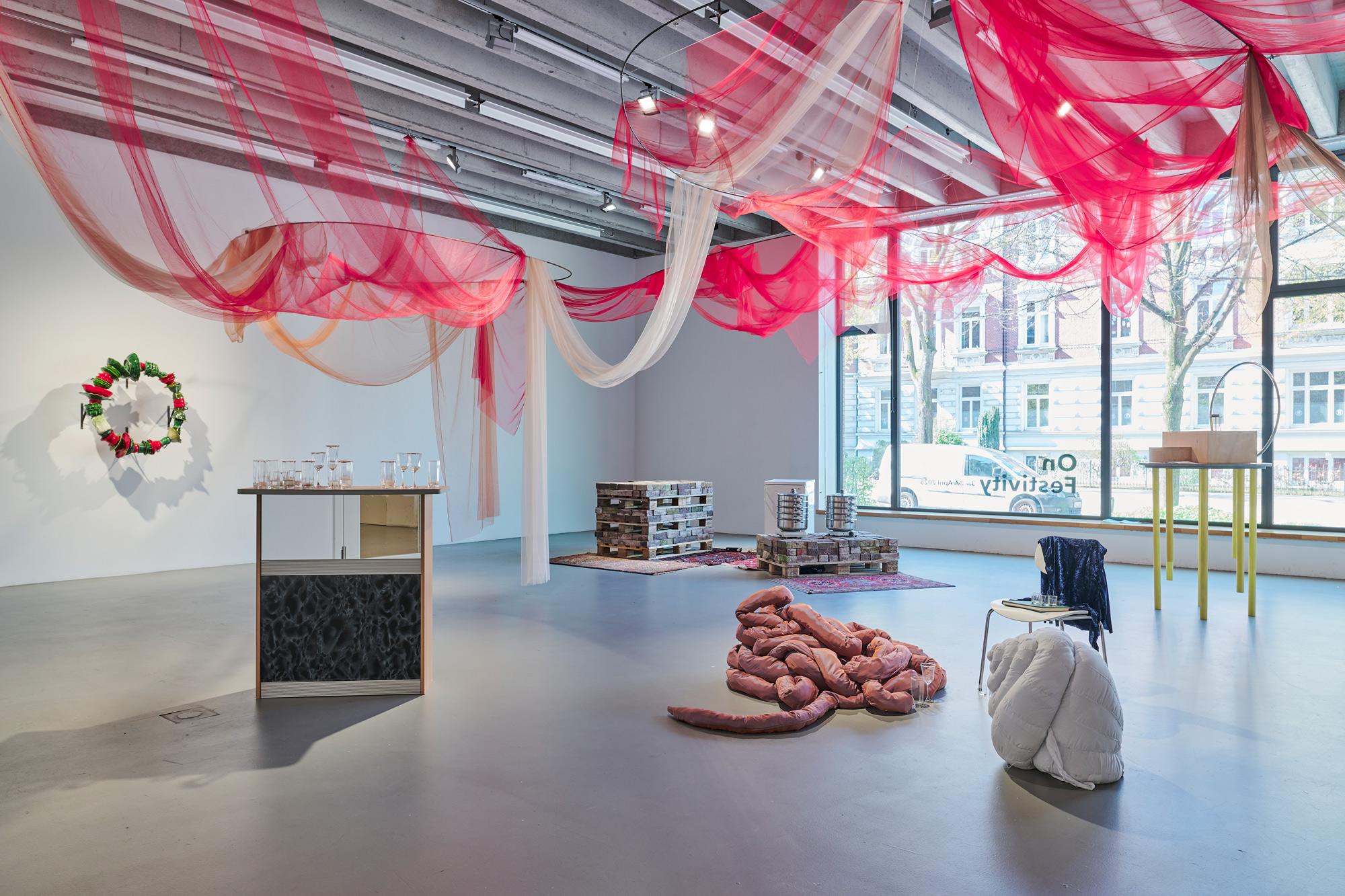 A bright exhibition space with large windows and an installation of pink and translucent fabric draped from the ceiling. Various objects and sculptures are arranged throughout the room, including stacked bricks, glass vessels on a table, and soft, organic-shaped elements on the floor.