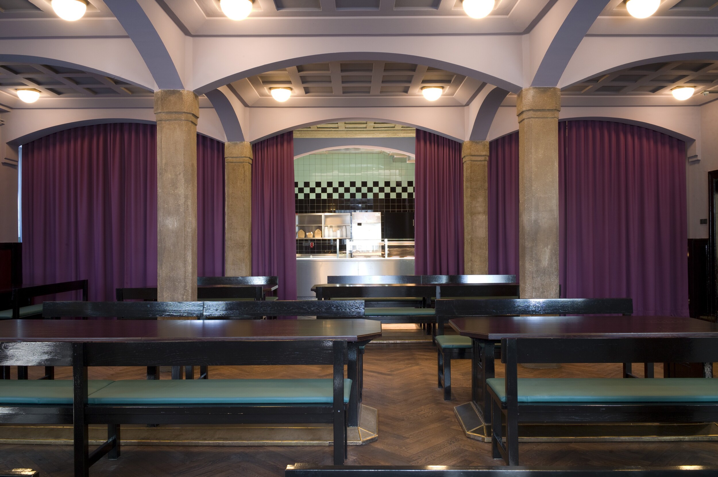 An empty dining hall with long, dark wooden benches and tables. Purple curtains hang between columns in the background, revealing an open kitchen area with mint-green and black tiles. The vaulted ceiling is lit by round ceiling lights.