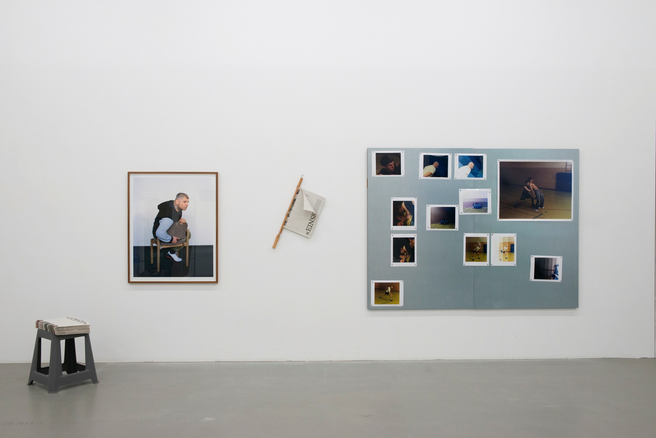 Gallery wall with a photograph of a seated man, a newspaper mounted at an angle, a collection of photographs on a gray background, and a small stool in front.