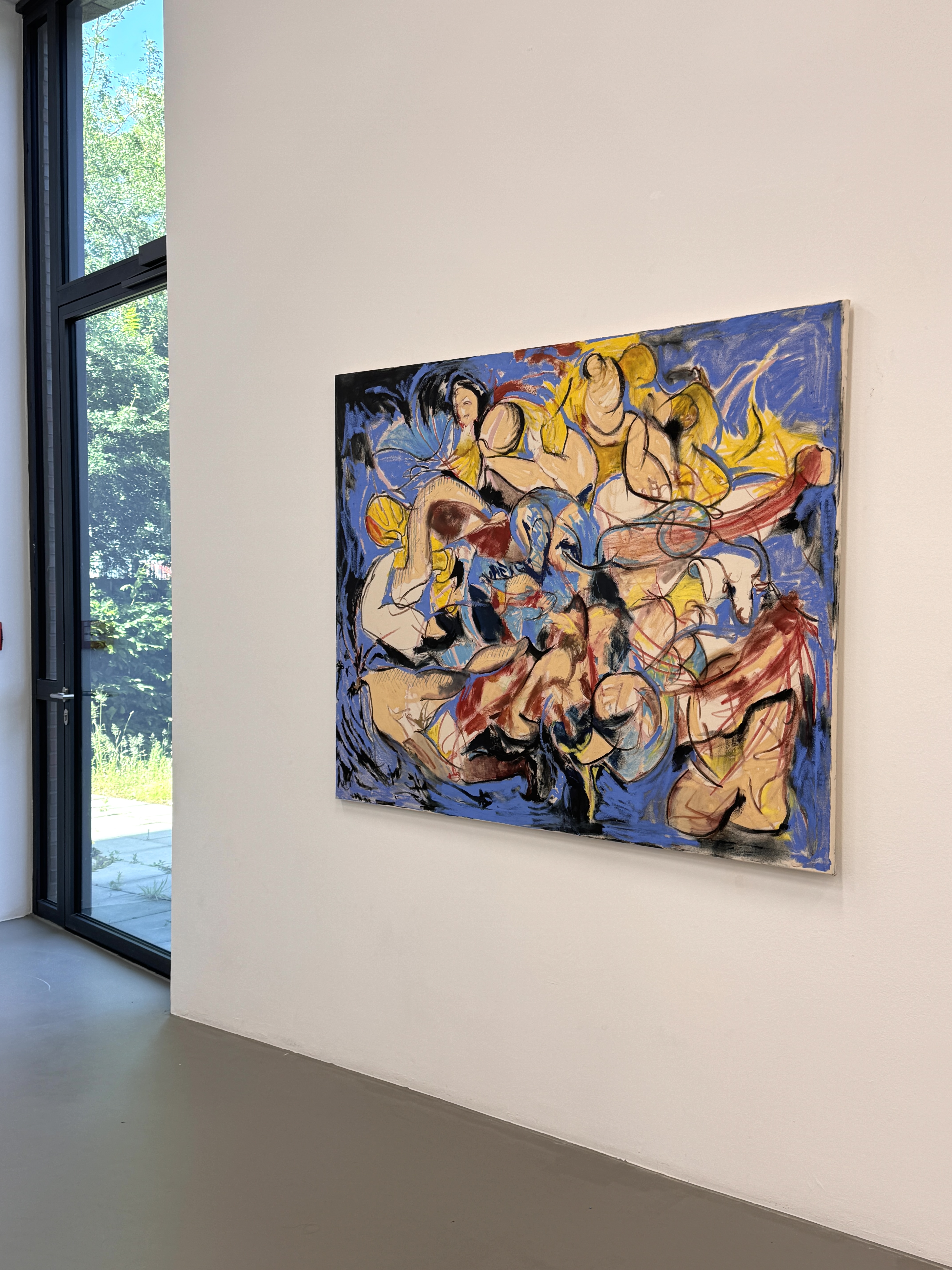 Painting with multiple abstract intertwined human figures in bold colors on a white wall in a room with a large window.