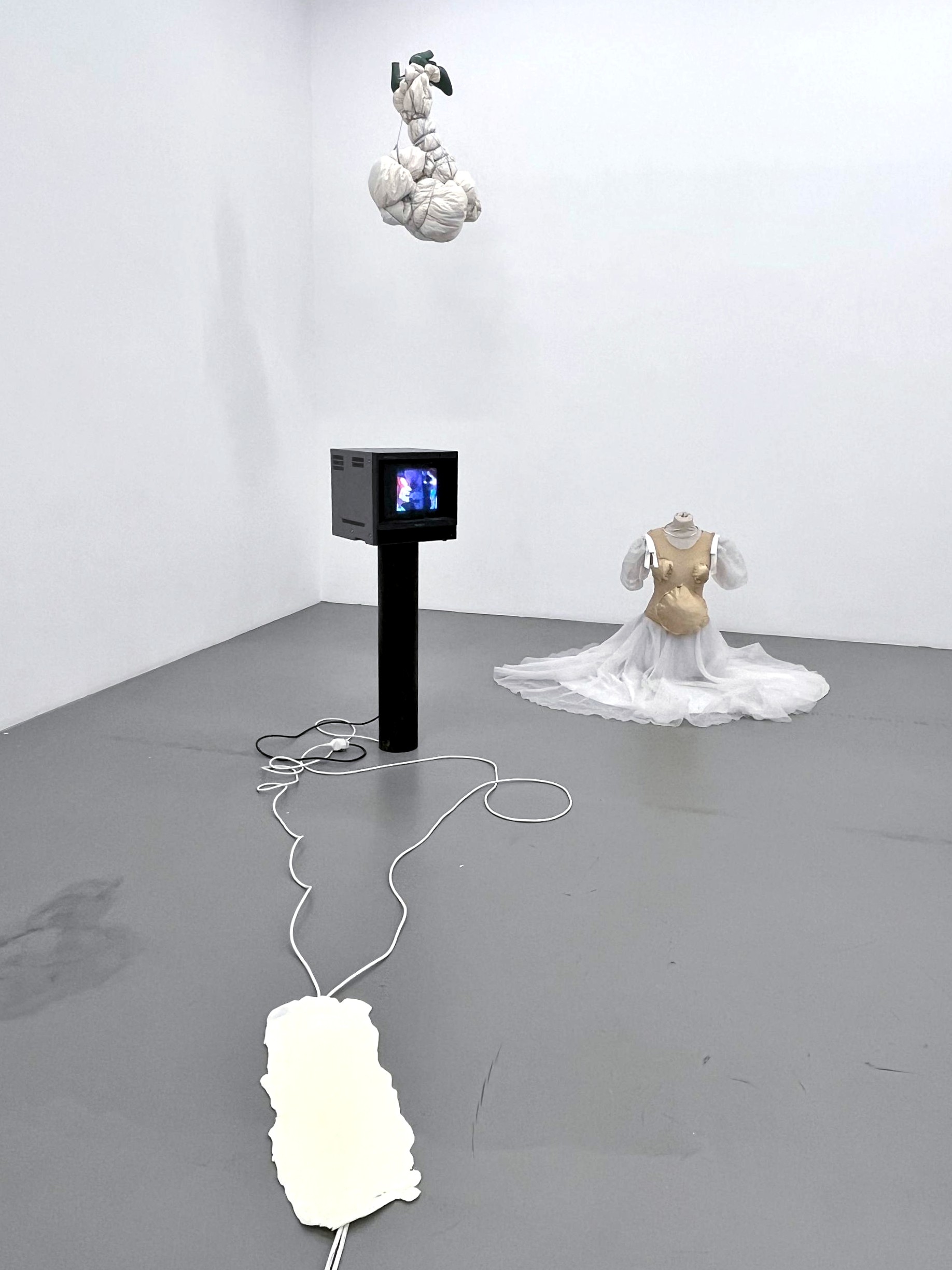 Black monitor on a black stand with cables on a gray floor, white fabric bundles hanging above, and a white dress with a beige bodice on the floor in a white gallery corner.