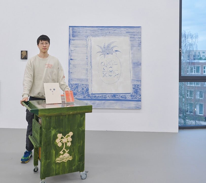 Kenneth Lin stands beside a green rolling cart decorated with a palm tree motif in front of a blue-and-white canvas featuring a stylized pineapple and circles.