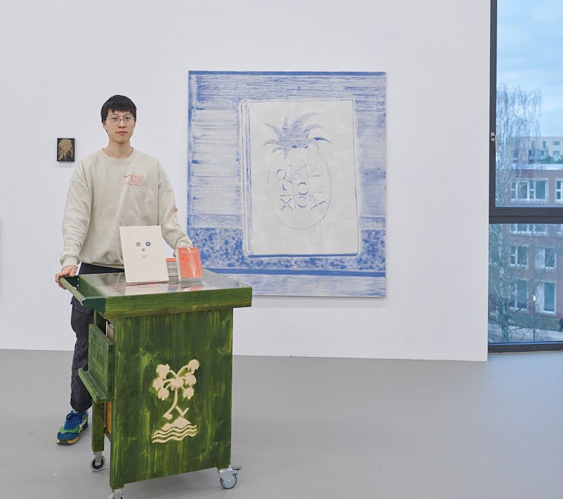 Kenneth Lin steht neben einem grünen Rolltisch mit Palmenmotiv vor einer blau-weißen Leinwand mit Ananas- und Kreismotiven