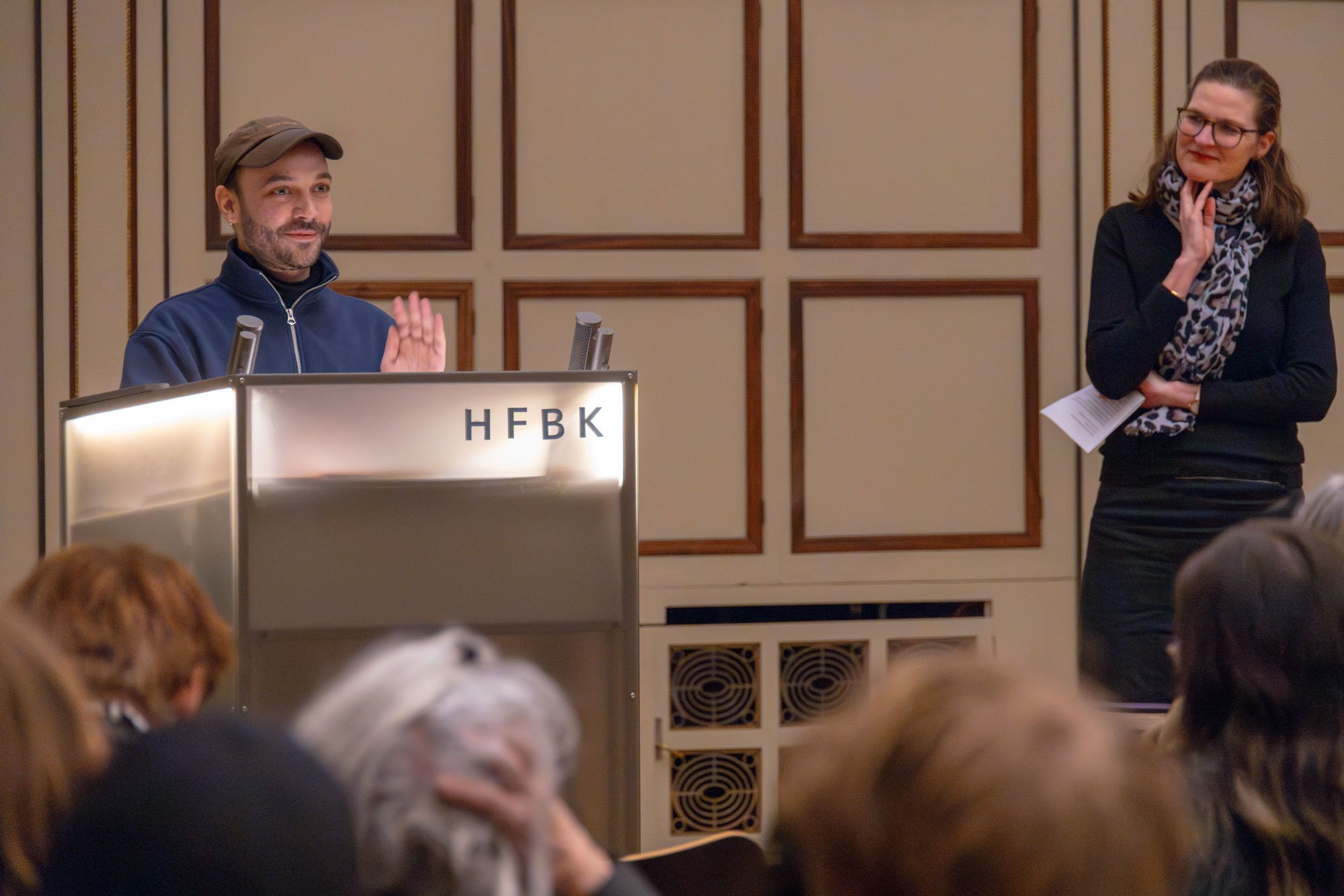 One person stands at a lectern labeled “HFBK,” speaking to an audience, while another person stands beside them listening with notes in hand. The heads of audience members appear blurred in the foreground, with a wood-paneled wall in the background.