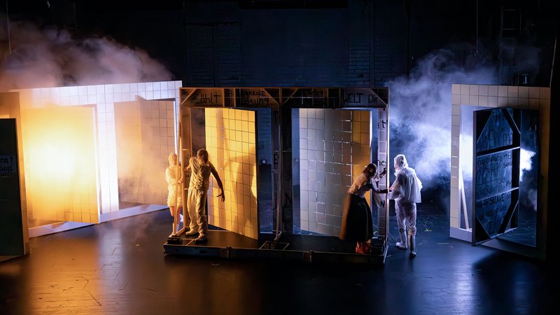 Theatrical scene featuring several walk-in, cube-shaped stage structures resembling tiled rooms. Warm and cool lighting, along with stage smoke, create a dramatic atmosphere. Several performers stand inside and between the structures, some appearing to open doors or interact with one another.