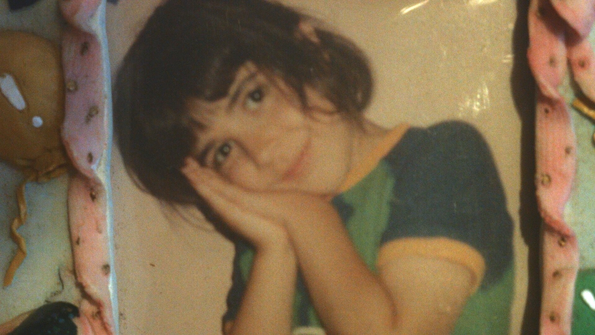 Child with dark hair wearing a green and blue t-shirt smiling and holding hands to face in a photo frame with pink decoration