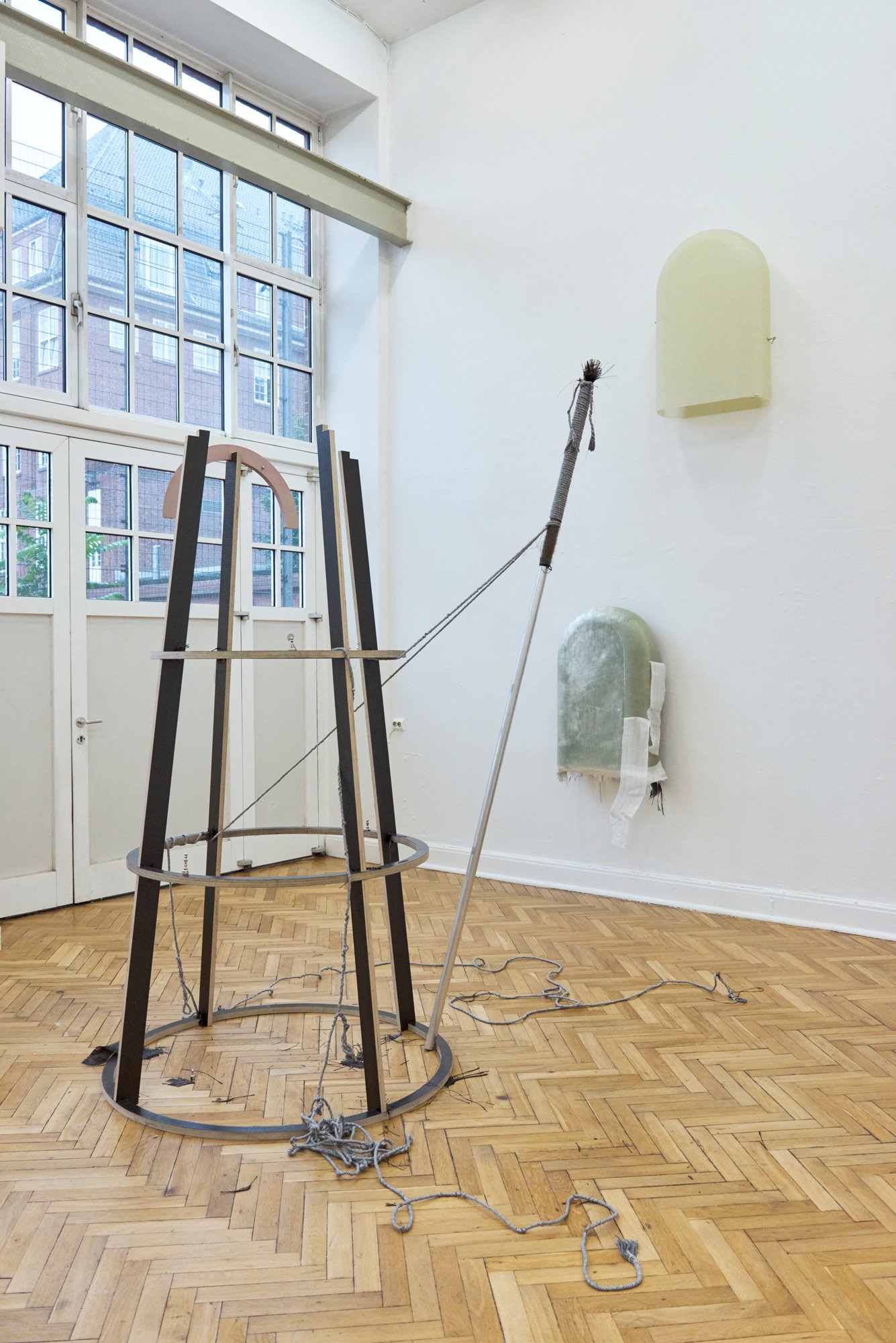 Exhibition view of a bright gallery space with parquet flooring and tall windows. At the center stands a sculptural construction made of metal rings, dark beams, and ropes. A long pole leans diagonally against the wall nearby. Two semi-arched wall-mounted objects are visible on the wall, one pale yellow and one greenish with a white fabric element.