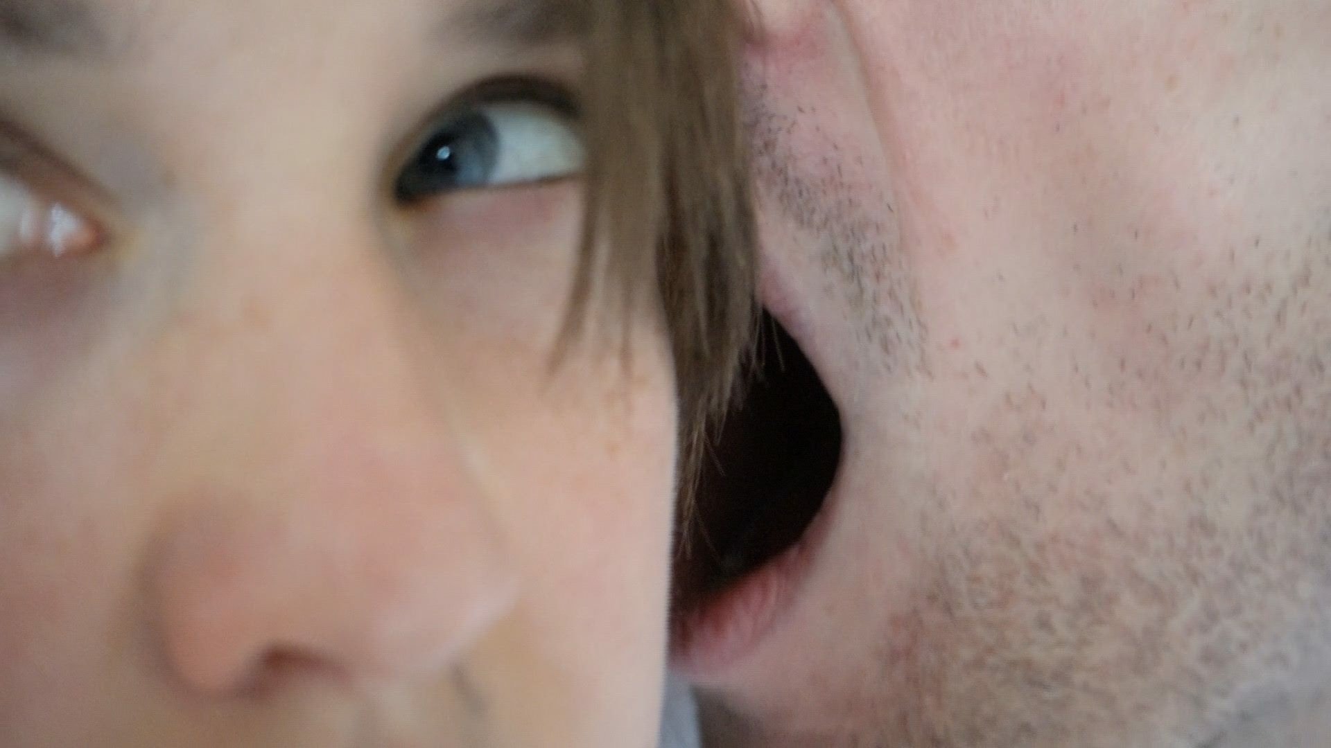 Extreme close-up of two faces: On the left, part of a person’s face is visible, including one eye looking to the side. On the right, another person with light stubble leans very close to the ear, as if whispering. The background is blurred.