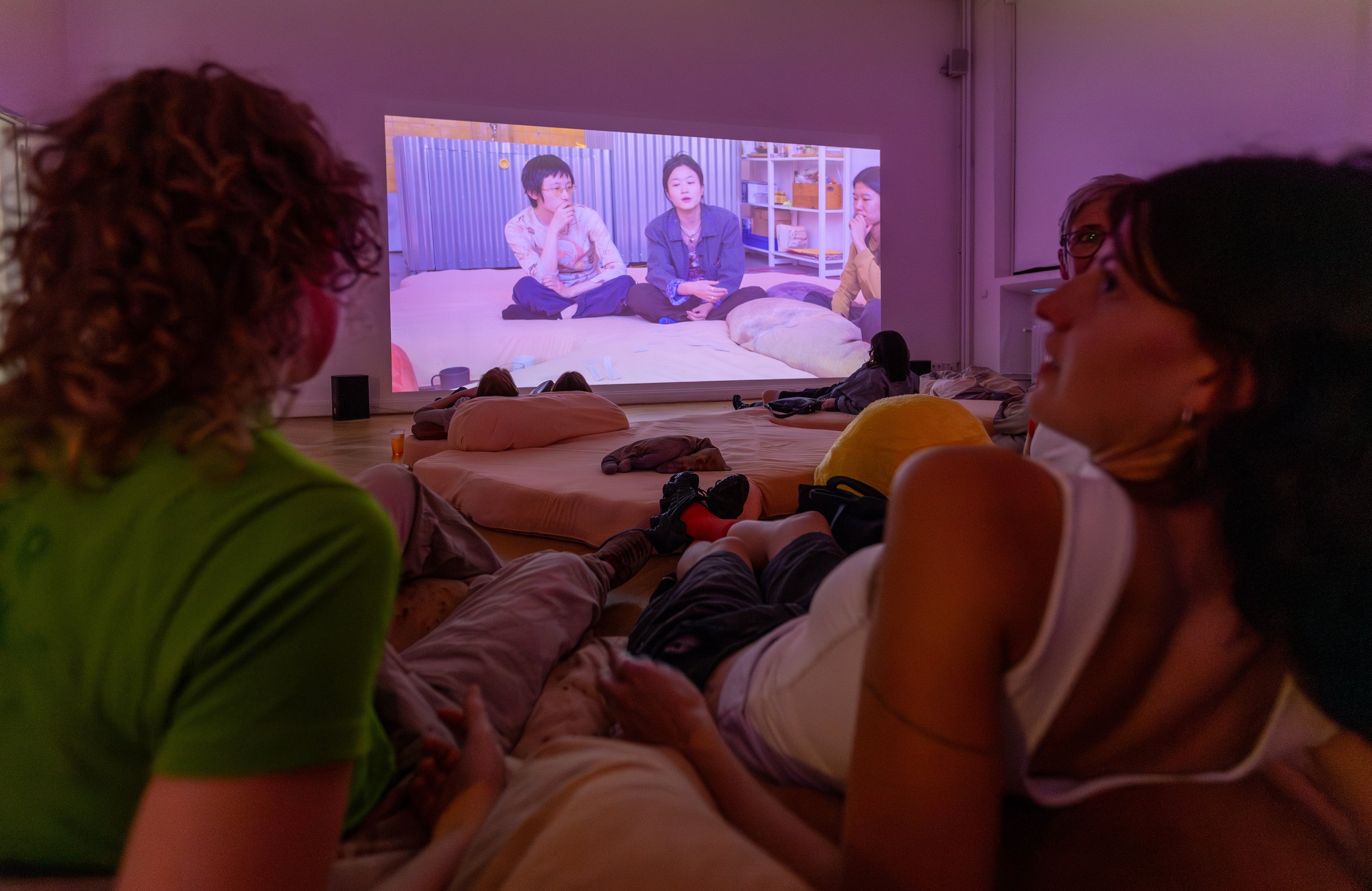 Zwei Besucherinnen liegen auf dem Boden zwischen Kissen. Das Foto wurde mit einem Blick zwischen die Schultern der beiden Personen aufgenommen. Der Blick geht auf eine Videoleinwand.