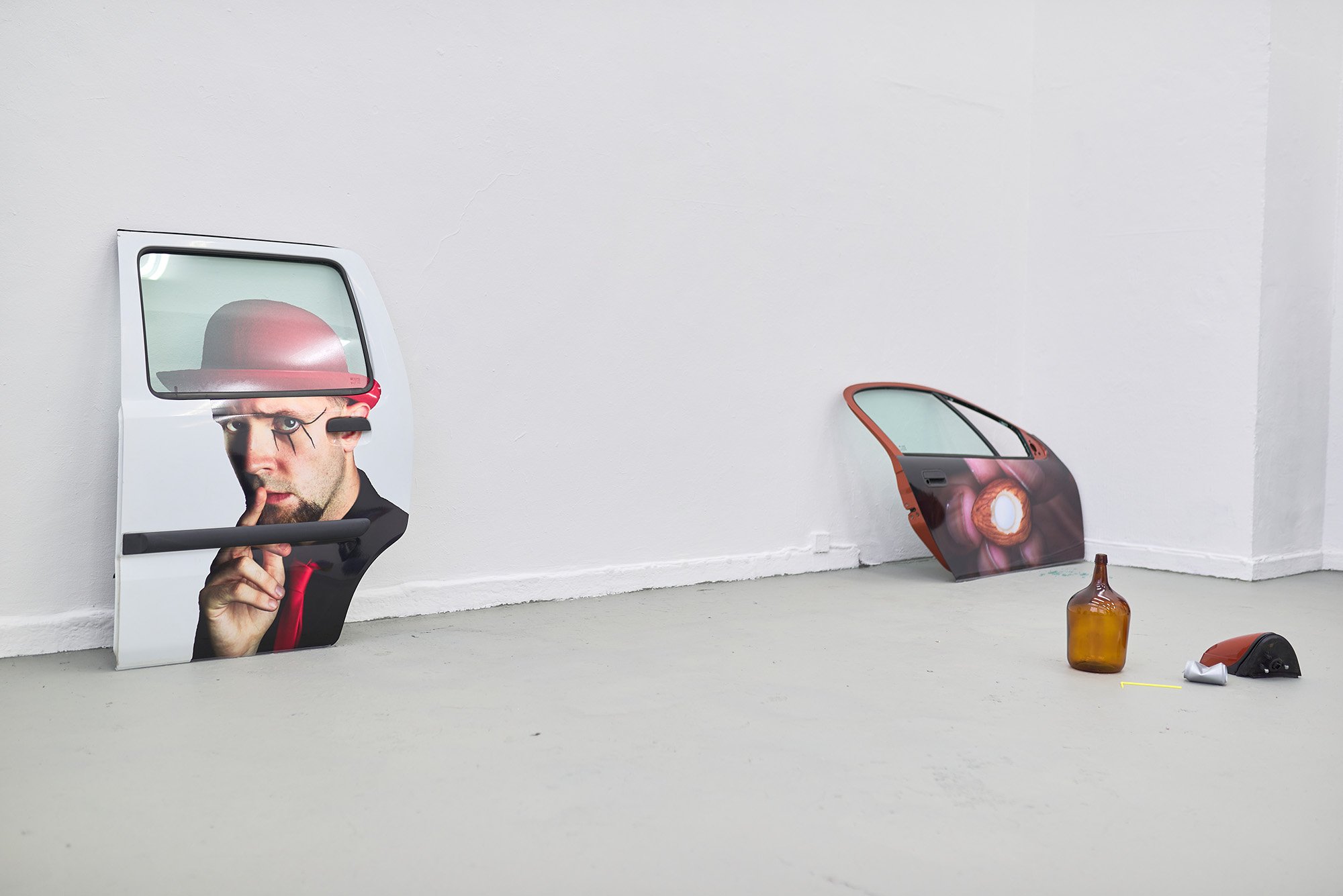 Installation in a white exhibition space featuring two painted car doors leaning against the wall. The left door shows a portrait of a man wearing a red hat and eye makeup, holding a finger to his lips. The right door depicts a large, detailed eye. On the floor in front are a brown glass bottle and a small dark object.