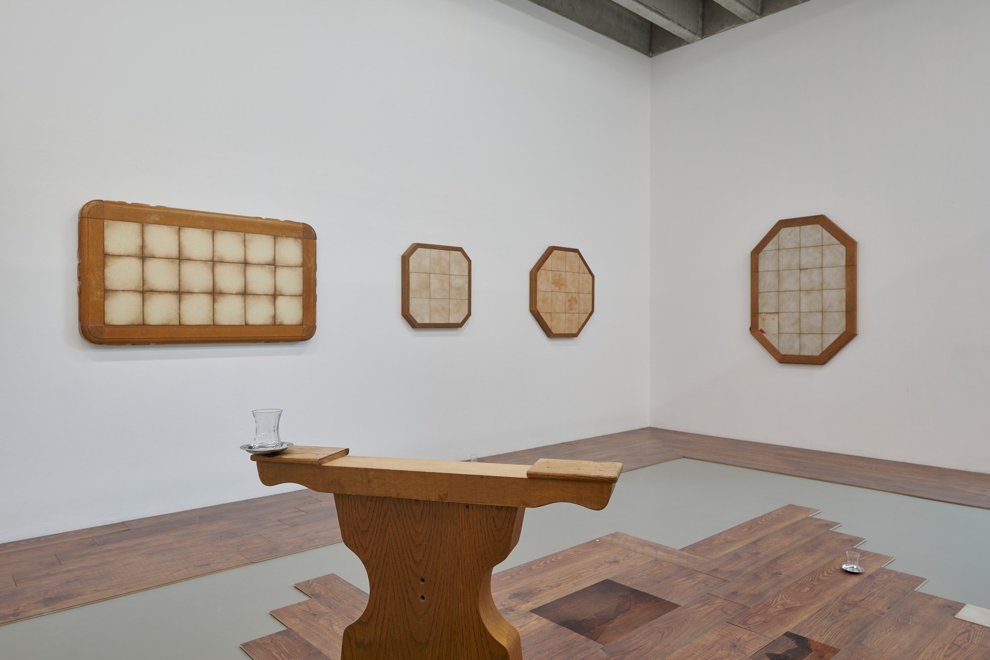 Installation view of an exhibition space with white walls. Several framed, geometric wall works with grid-like surfaces are mounted on the walls. In the room stands a wooden, podium-like object holding a glass cup. The floor is partly covered with wooden panels framing a recessed area.