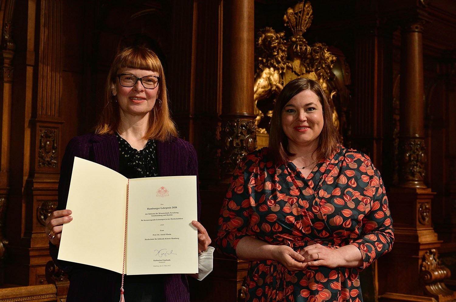 Two women stand in an ornate historic hall with dark wood paneling and a golden statue in the background. One of them holds an open certificate and presents it toward the camera. Both are smiling and looking forward.