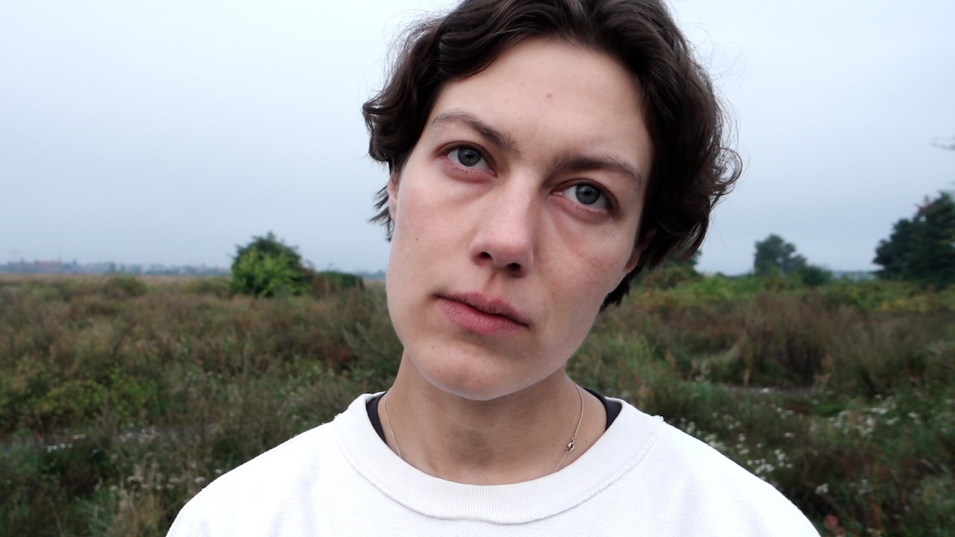 Close-up of a person with short dark hair looking directly into the camera. They are standing outdoors in an open, lightly overgrown landscape under a gray, overcast sky. Their expression appears calm and contemplative.