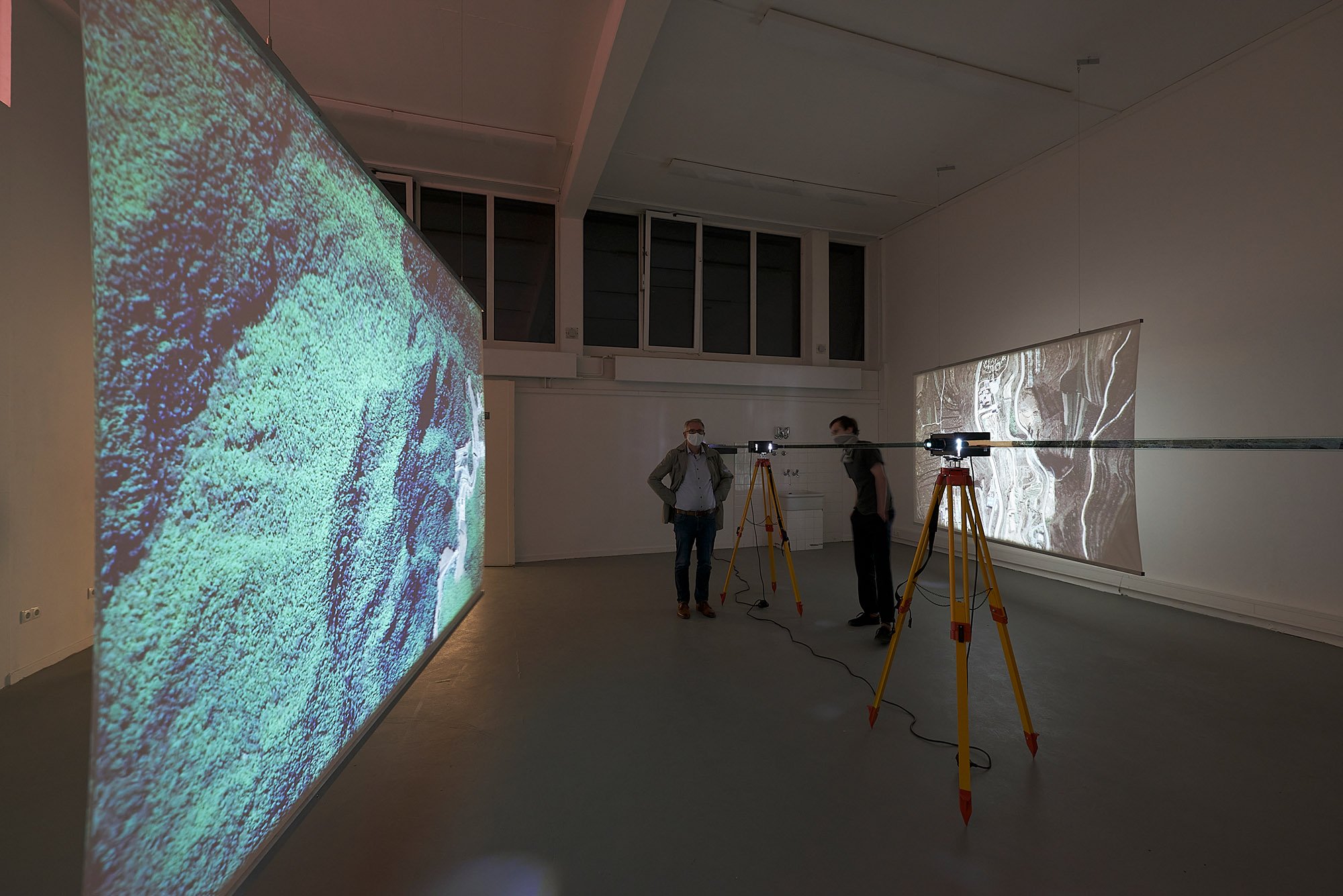 Exhibition view in a dimly lit room with two large projections on opposite walls. In the center, two projectors mounted on yellow tripods are connected by a glass panel. Two people stand nearby, observing the installation.