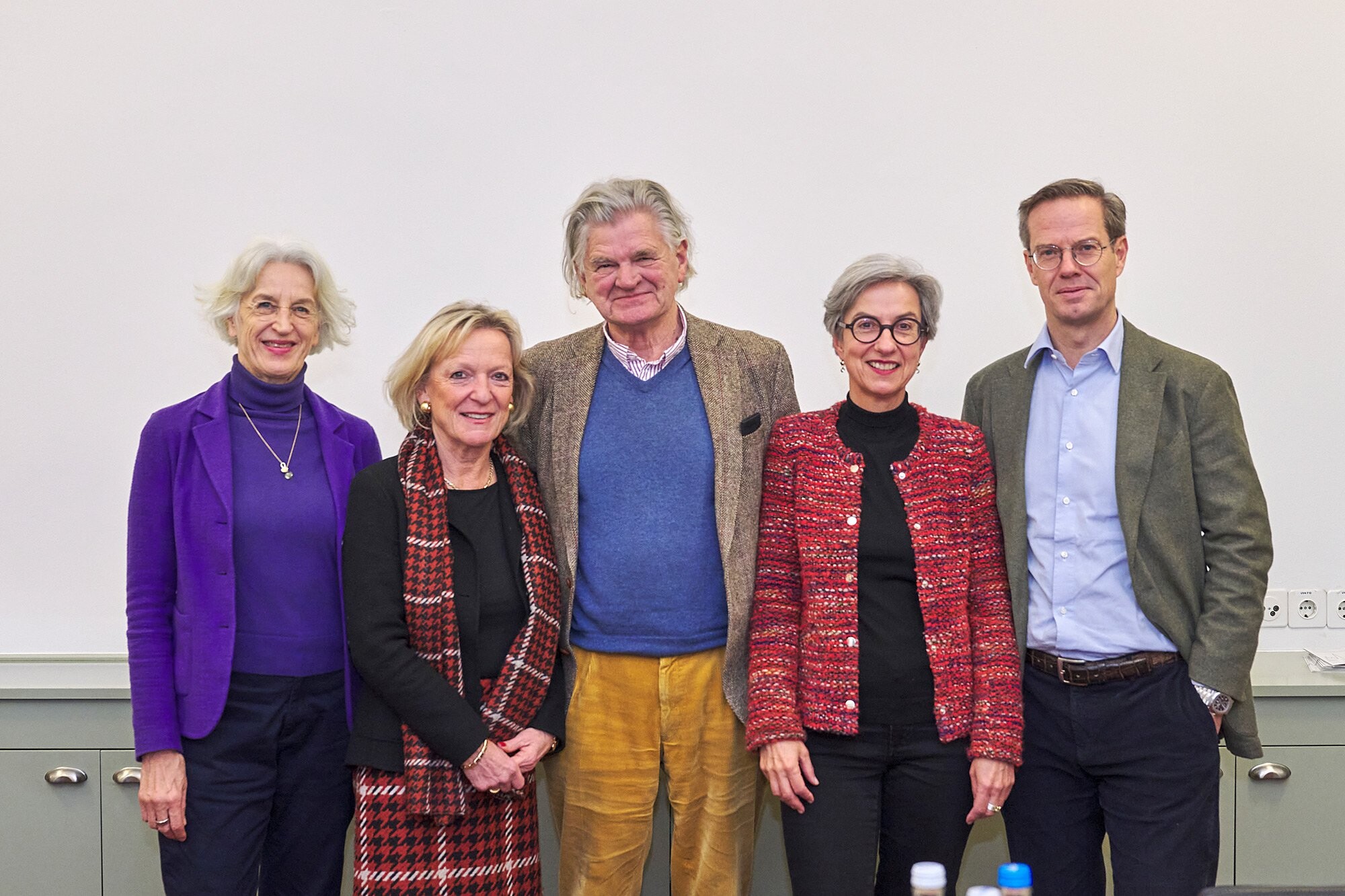Five people stand side by side for a group photo indoors in front of a light-colored wall. Three women are on the left, a man is in the center, and another man is on the right. They are all smiling at the camera and wearing formal to semi-formal clothing in different colors.